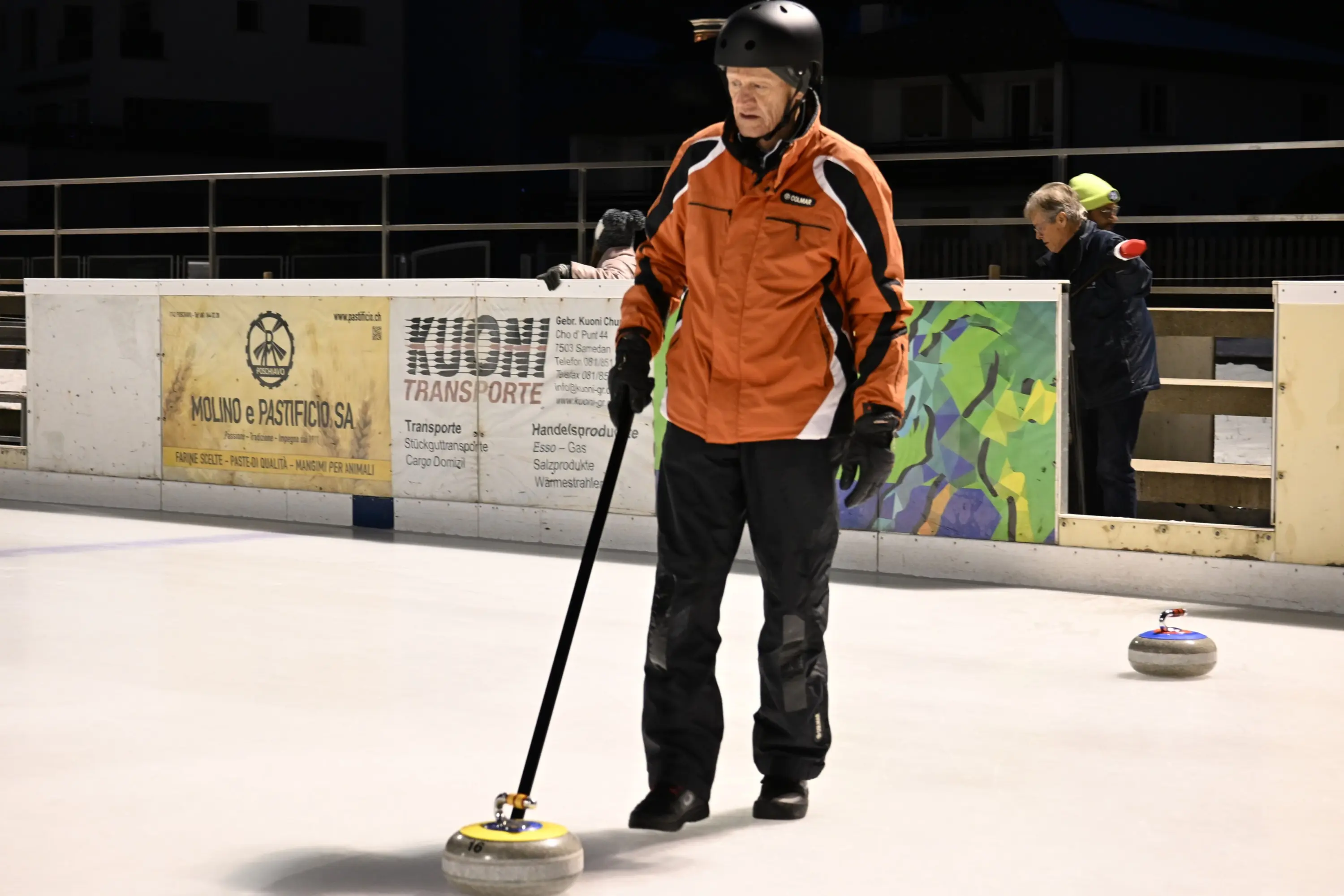 Un uomo anziano in giacca arancione e casco nero sta praticando curling, mentre si prepara a lanciare una pietra sul ghiaccio. In background, altre persone osservano.