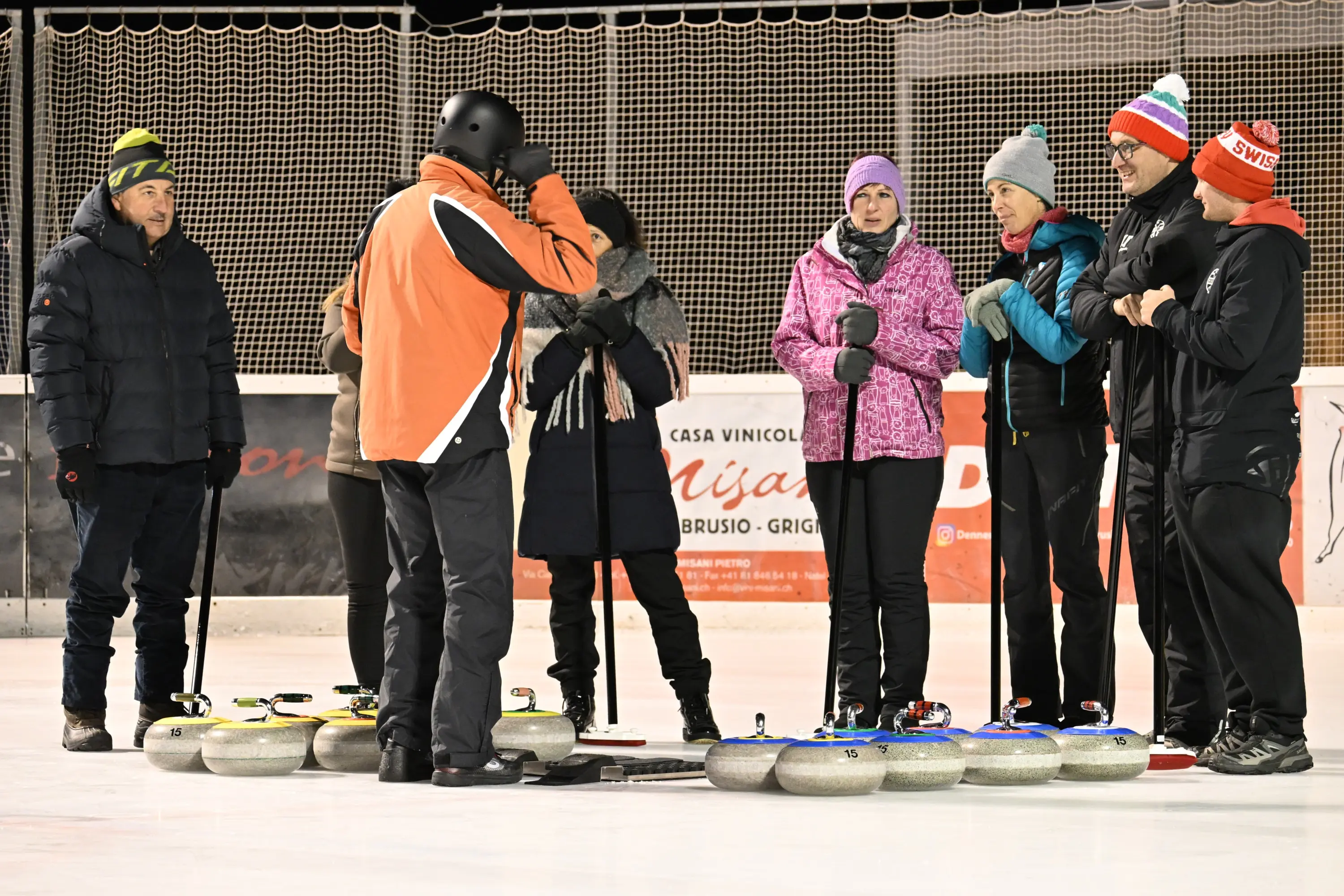 Un gruppo di sei persone discute ed esamina le pietre da curling su una pista ghiacciata. Alcuni indossano giacche calde e cappelli colorati. La scena