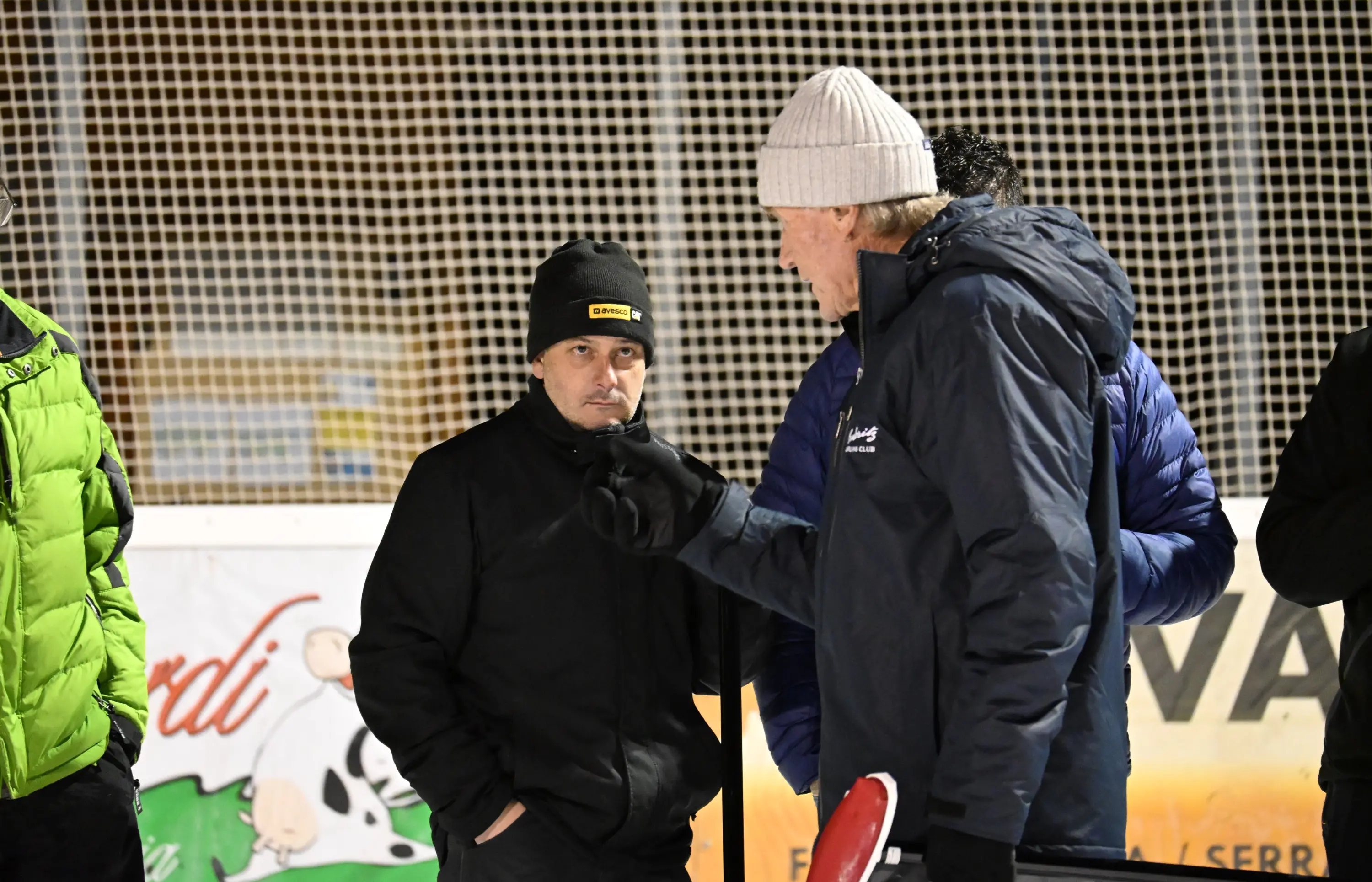 Due uomini conversano al freddo, indossando giacche pesanti e berretti. Uno sembra concentrato, mentre l'altro gesticola mentre parla. Sullo sfondo