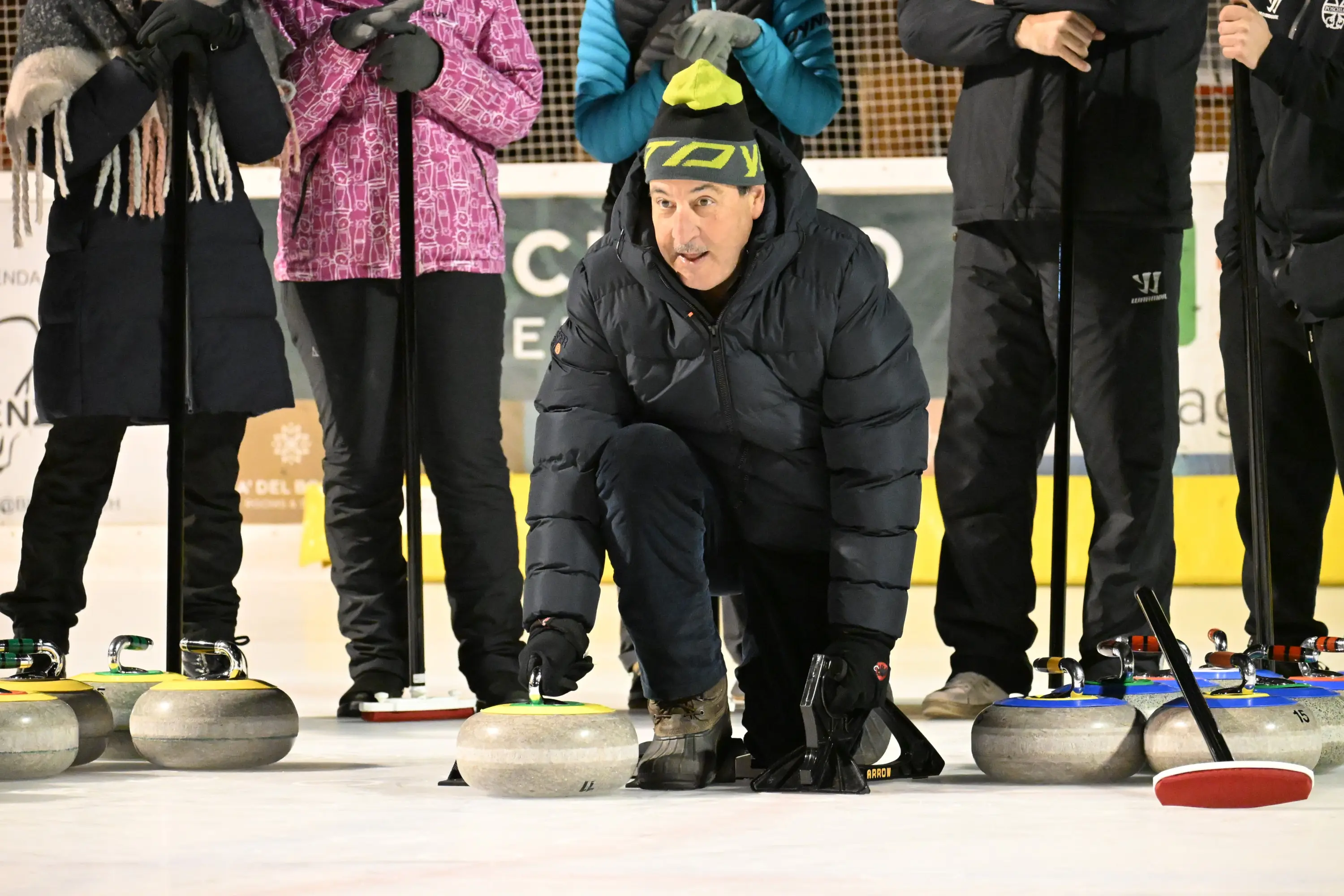Un uomo in abbigliamento invernale è in ginocchio su un campo di curling, pronto a lanciare una pietra. Sullo sfondo, altre persone osservano vest