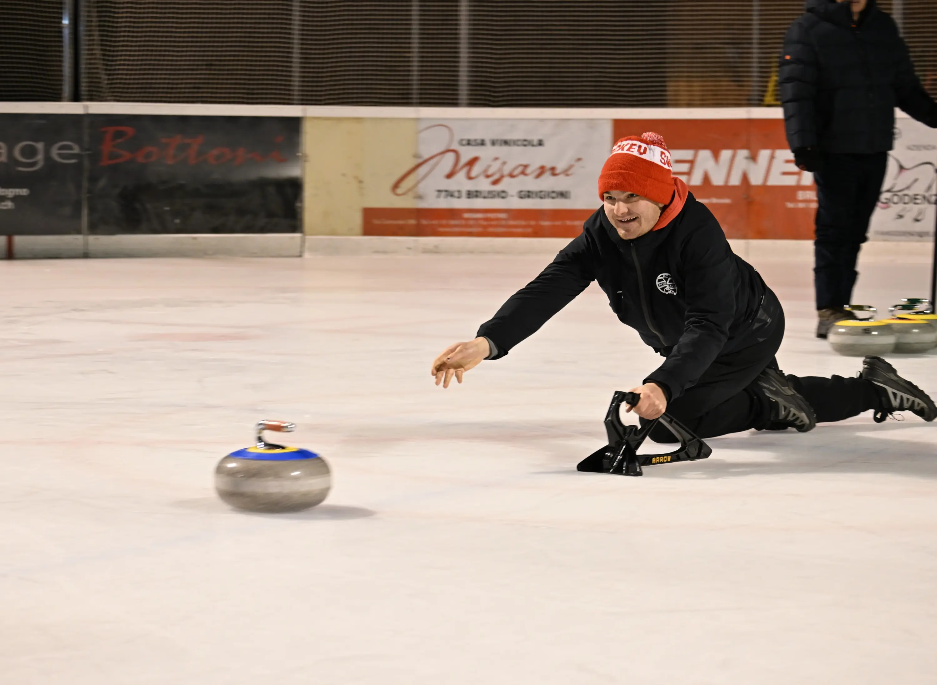 Un uomo in tuta nera e berretto rosso si allunga sul ghiaccio mentre lancia una pietra da curling. Un'altra persona è visibile sullo sfondo.