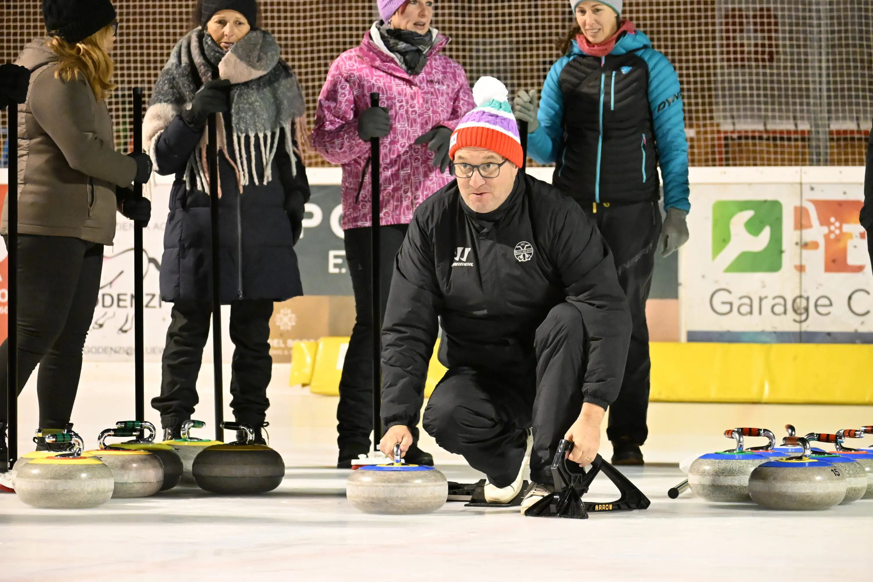 Un coach di curling è accovacciato sul ghiaccio mentre sistema una pietra da curling. Alle sue spalle, un gruppo di persone osserva, indossando abbigliamento in