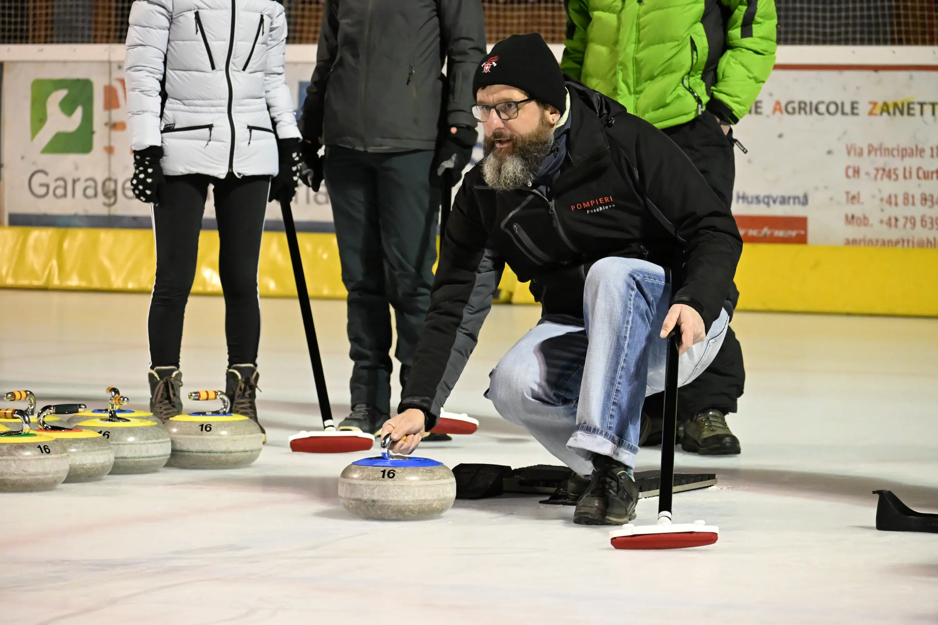 Un uomo con barba grigia e occhiali si inginocchia su una superficie di ghiaccio mentre posiziona una pietra da curling. Indossa un berretto e una giacca n