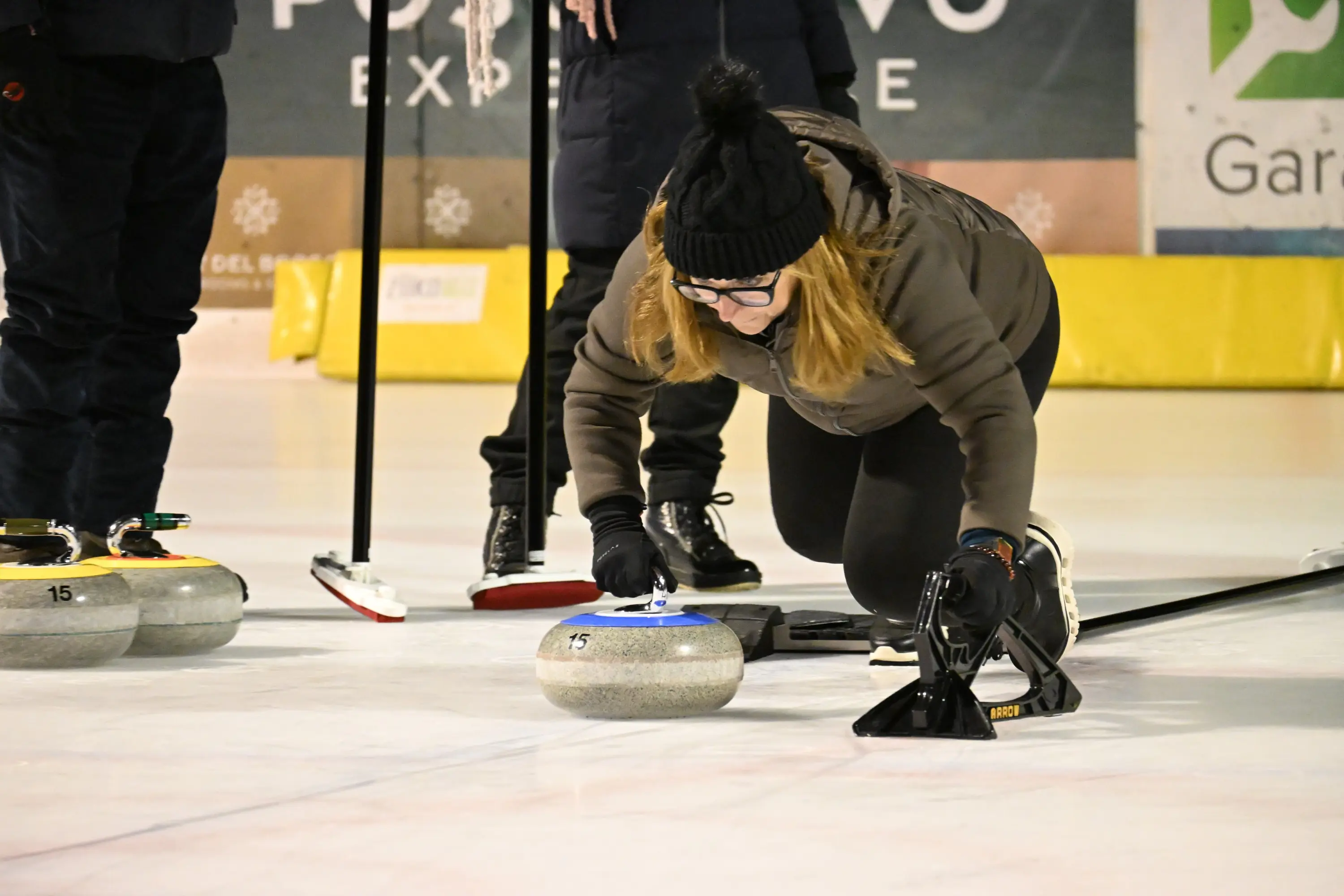 Una donna si piega in avanti mentre lancia una pietra da curling su una pista di ghiaccio. Indossa un cappello e guanti, concentrata nel suo gesto. Sul ghiaccio