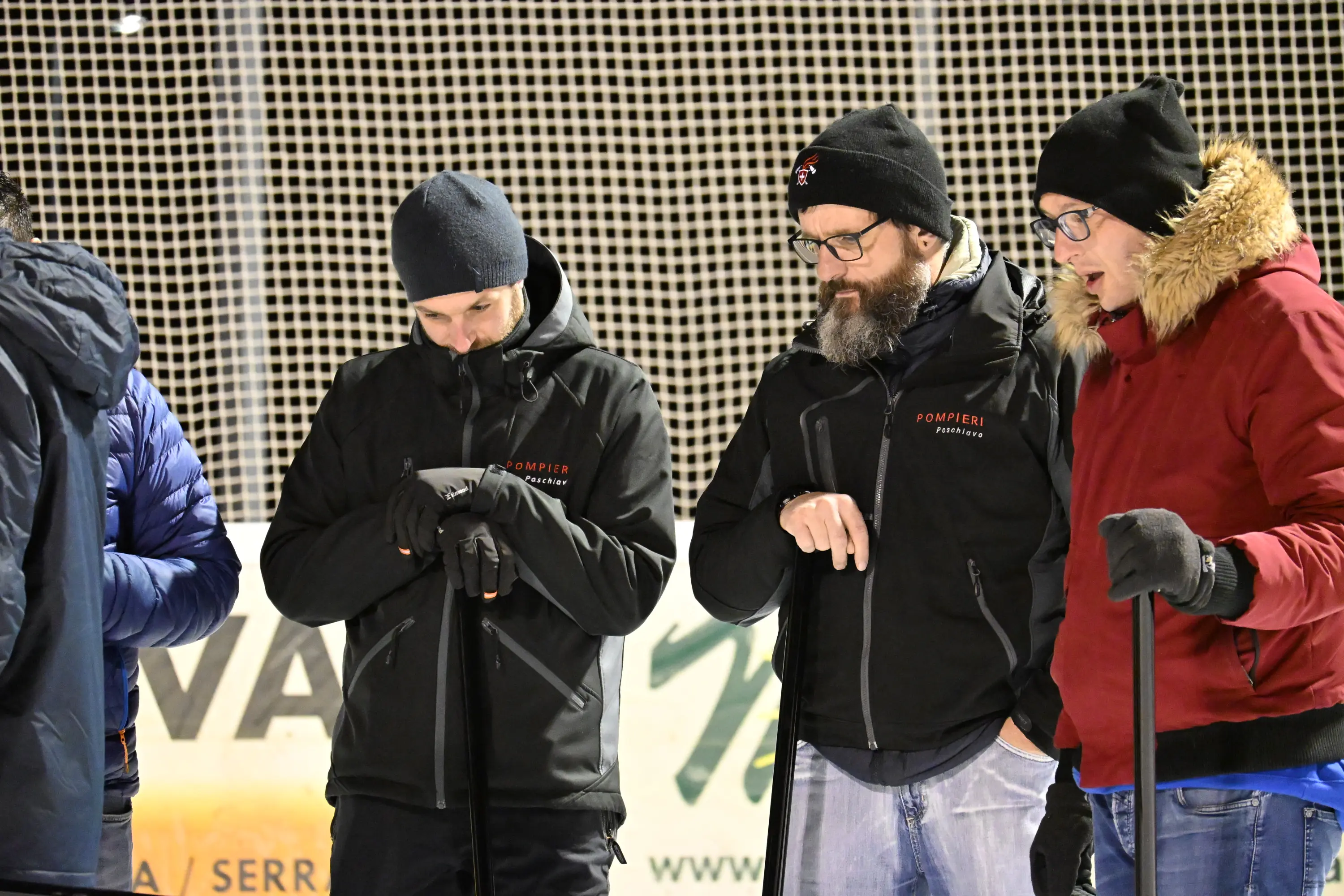 Tre uomini in abbigliamento invernale, uno con barba e occhiali, stanno osservando mentre tengono in mano dei bastoni. Si trovano su un campo da ghiaccio