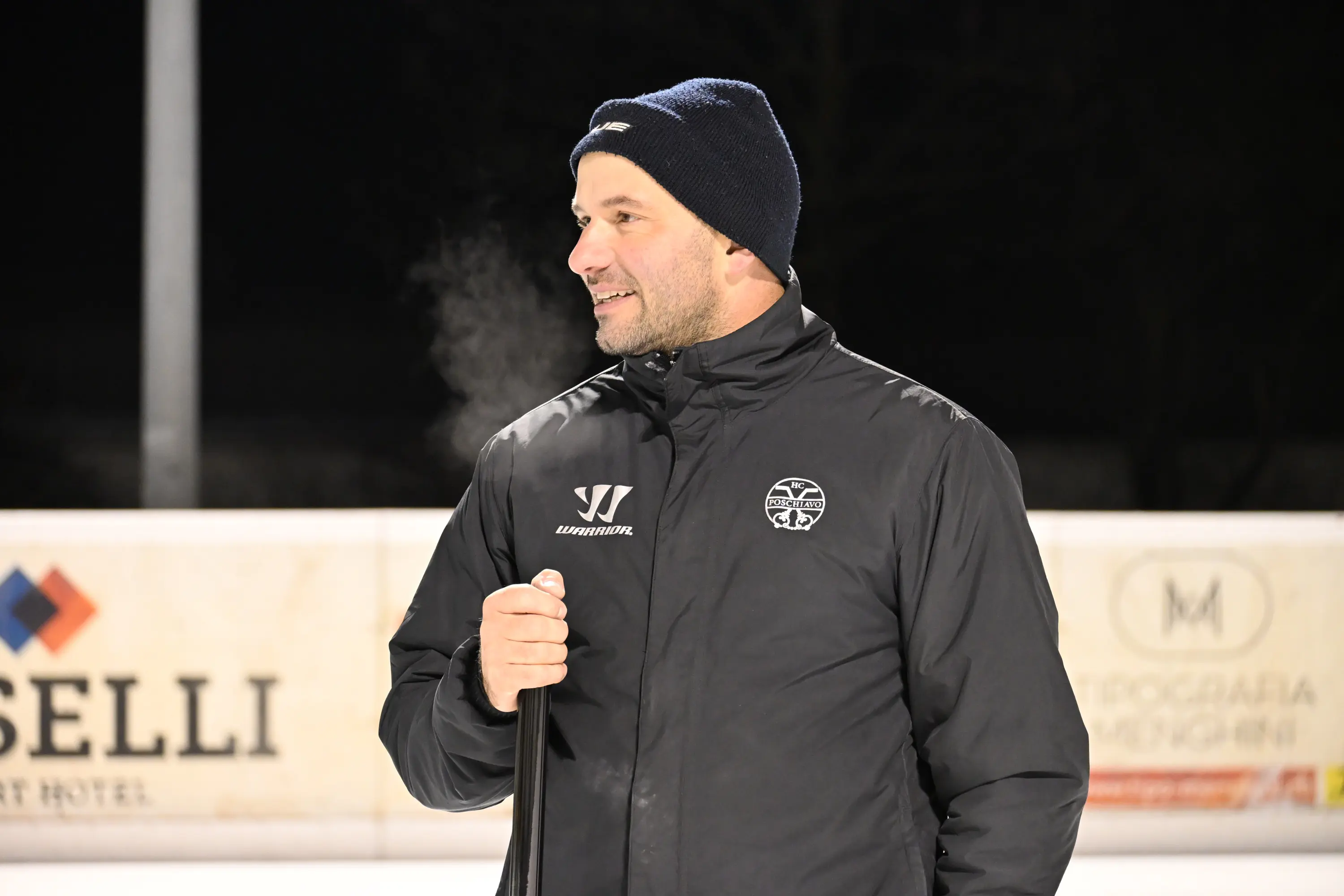 Uomo sorridente in abbigliamento invernale, con giacca nera e berretto, che tiene un bastone da hockey su ghiaccio. Si nota vapore che esce
