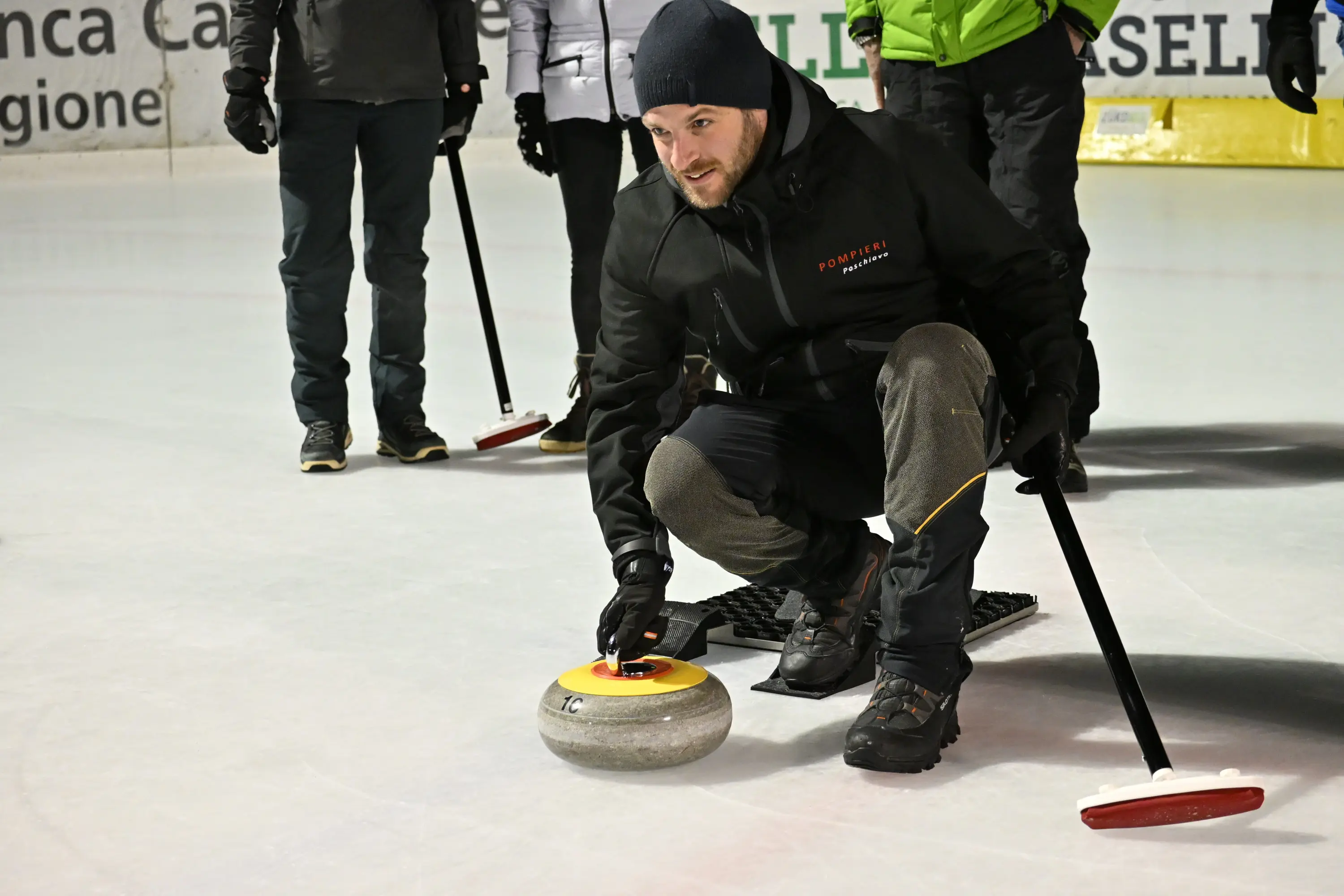 Un uomo in abbigliamento sportivo si sta preparando a lanciare un disco di curling su un campo di ghiaccio. Indossa una giacca nera e pantaloni grigi,