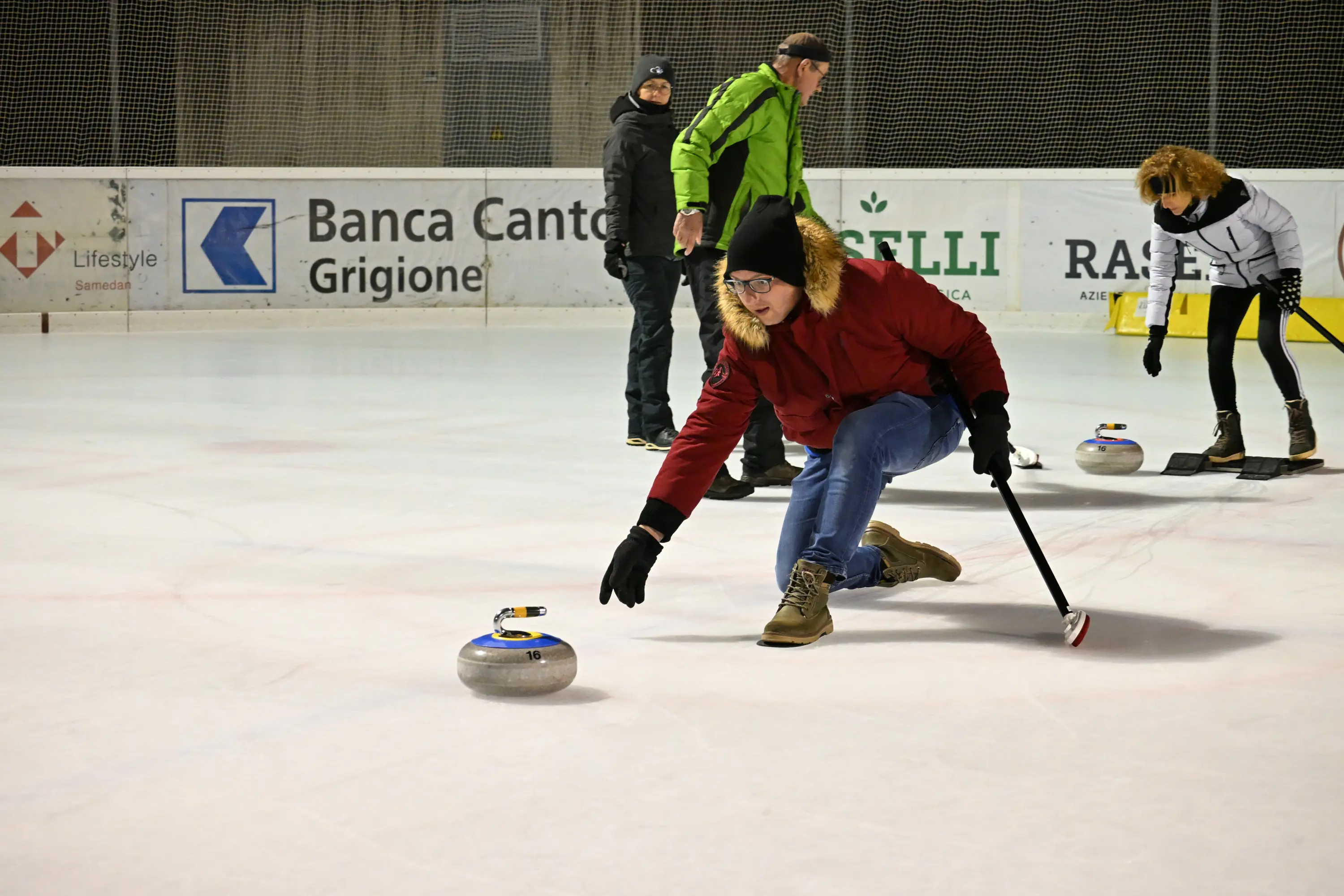 Una persona con giacca rossa lancia una pietra da curling su una pista di ghiaccio, mentre altre tre persone osservano.