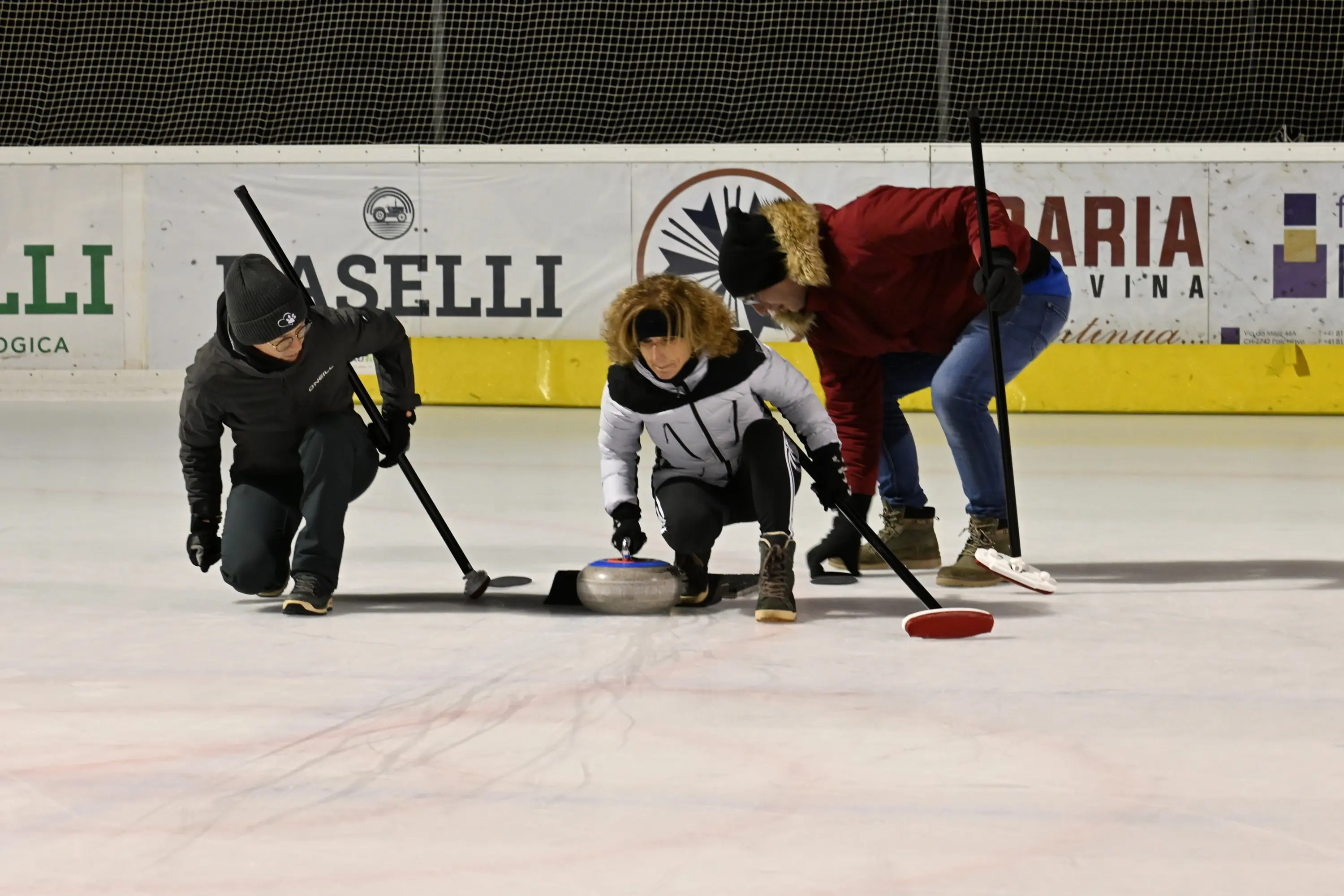Tre persone si chinano su un campo di curling ghiacciato, mentre si preparano a lanciare una pietra. Due partecipanti hanno una scopa in mano e indossano ab