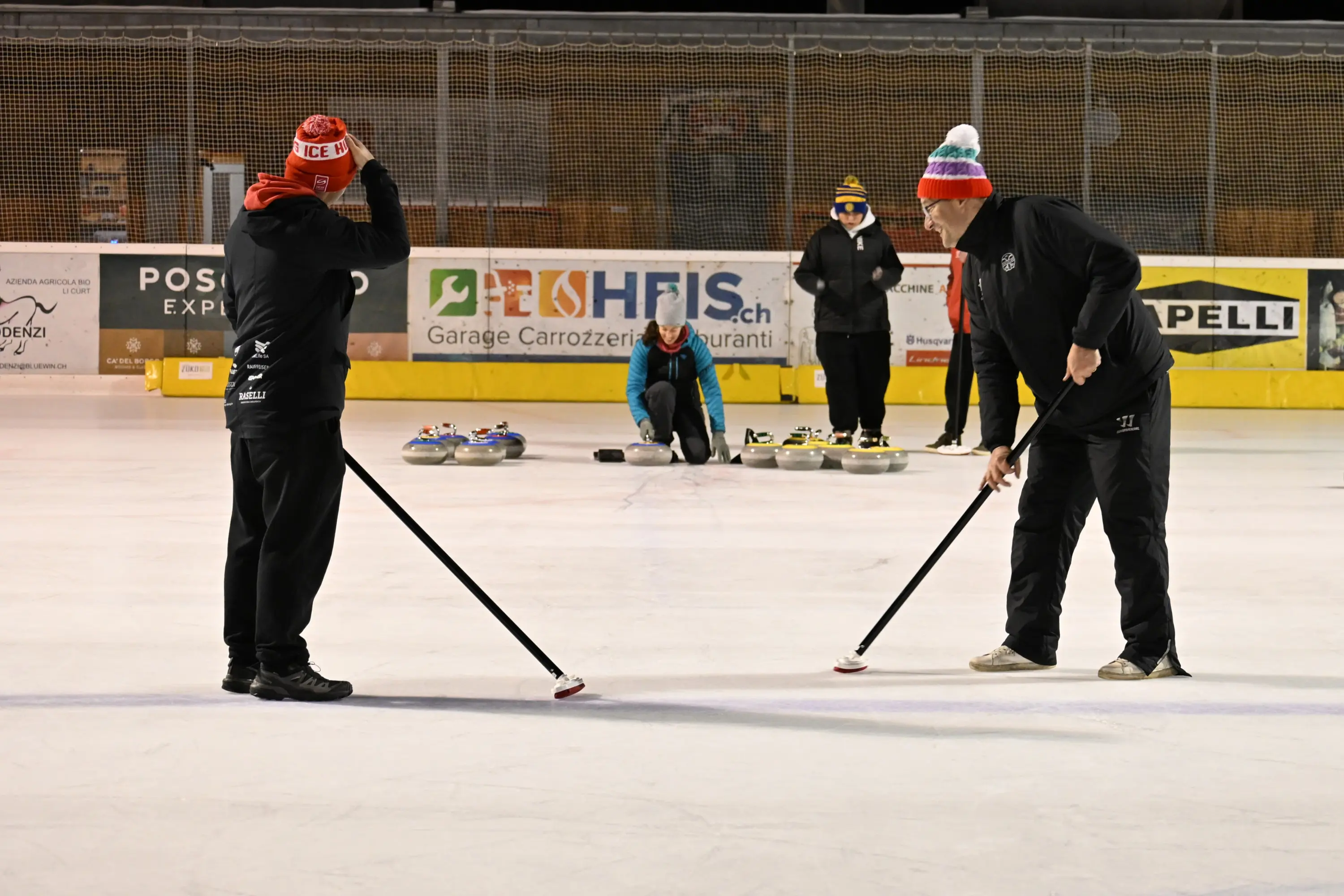 Due giocatori di curling si preparano a lanciare un sasso su una pista di ghiaccio. Indossano cappelli rossi e neri. Sullo sfondo, altri due