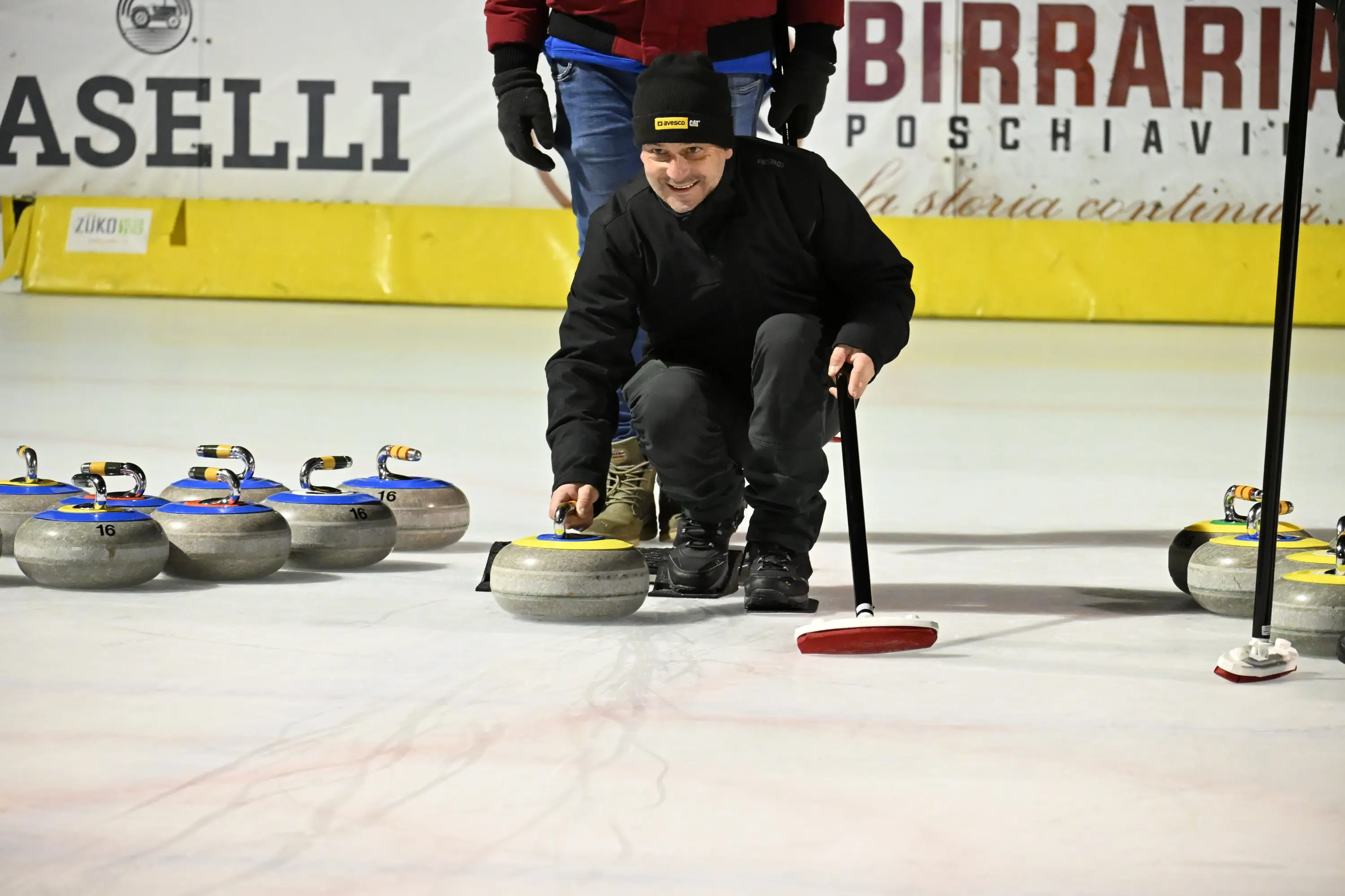 Un uomo in abbigliamento scuro si china per posizionare una pietra da curling sul ghiaccio, circondato da altre pietre. Il ghiaccio presenta segni di gioco