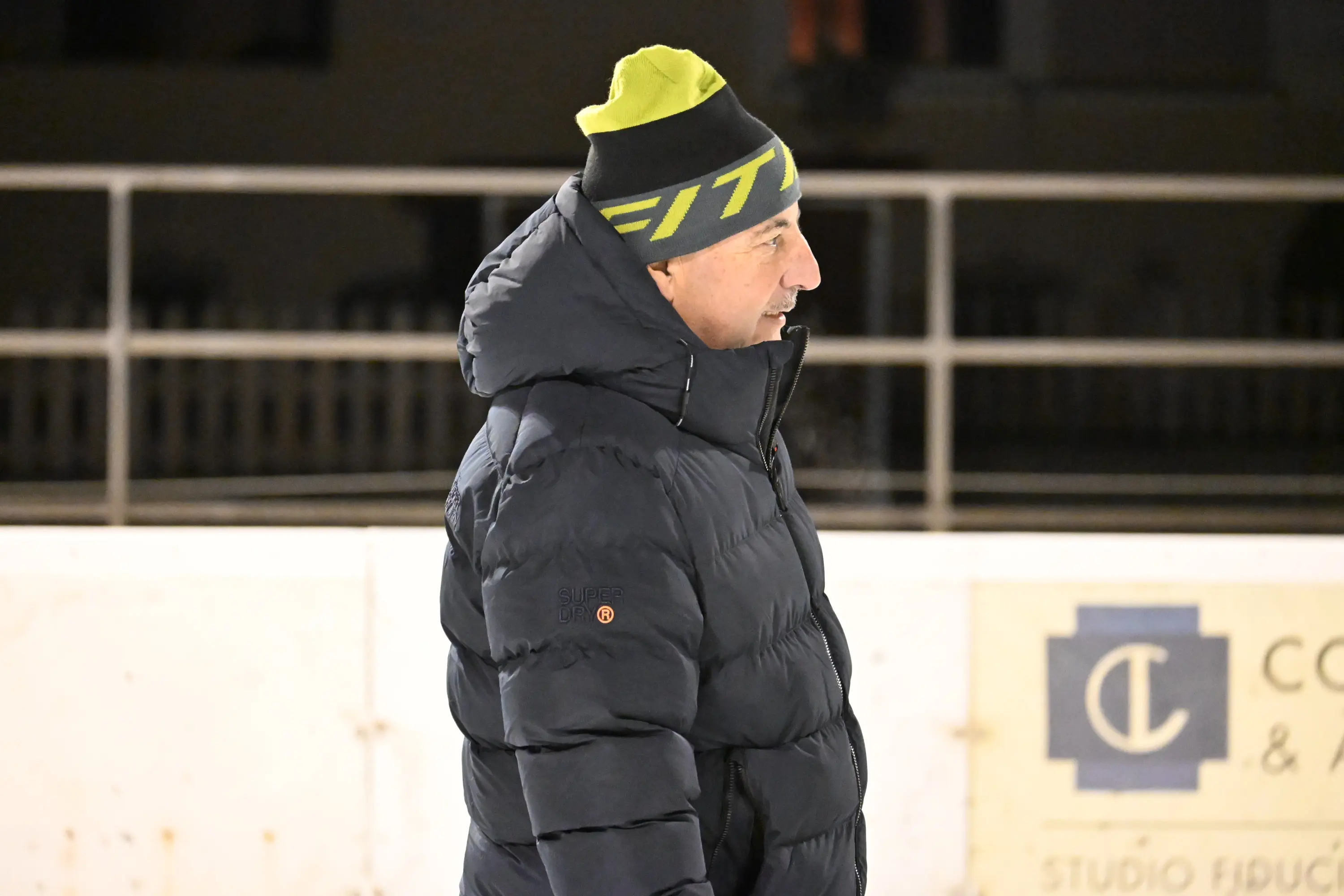 Uomo con cappotto pesante e berretto colorato, osserva da un campo di ghiaccio. Sullo sfondo, una recinzione bianca e un edificio poco illuminato.