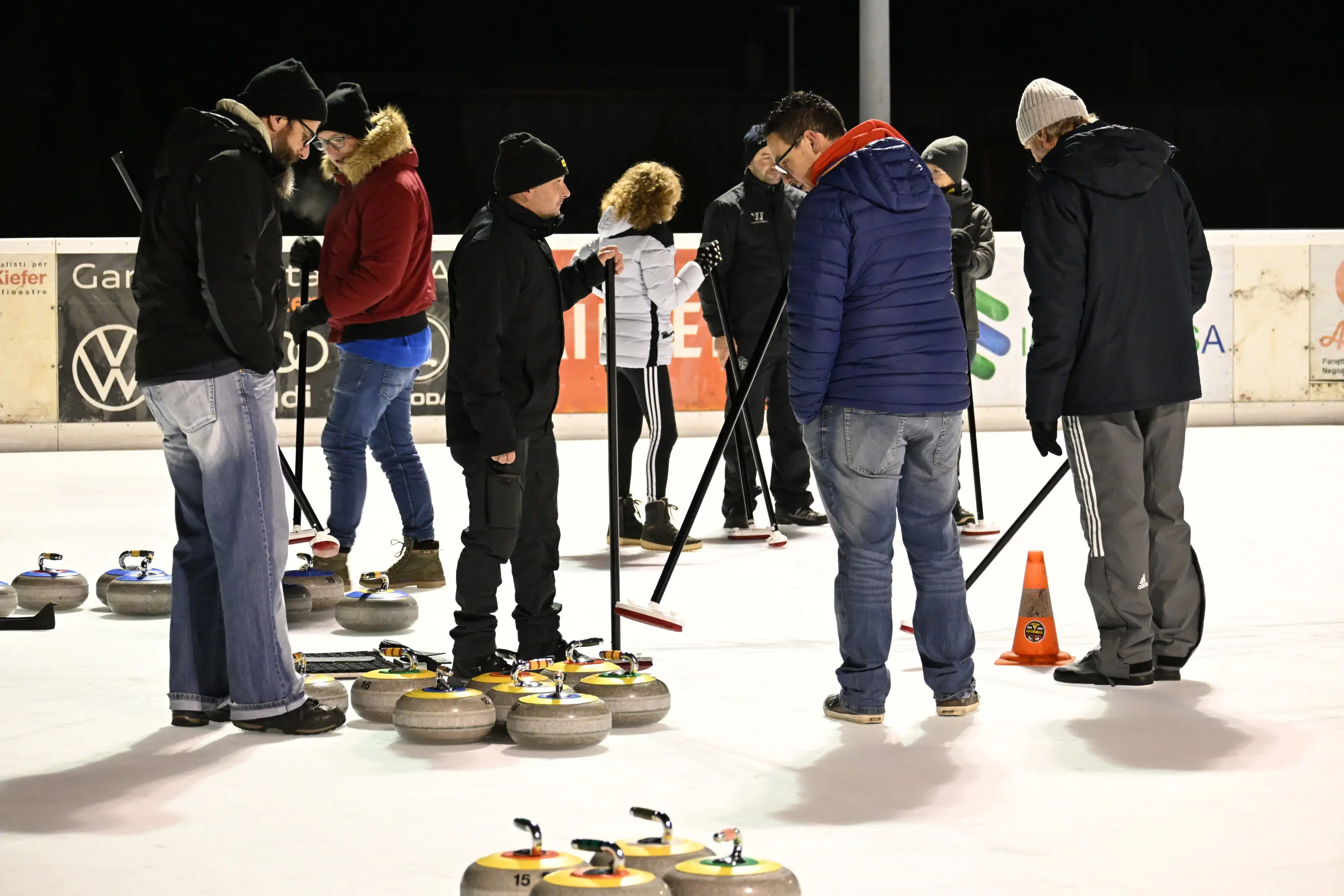 Gruppo di persone che si riunisce su un campo di curling al chiaro di luna. Alcuni indossano giacche e cappelli, mentre altri stanno discutendo e osservando
