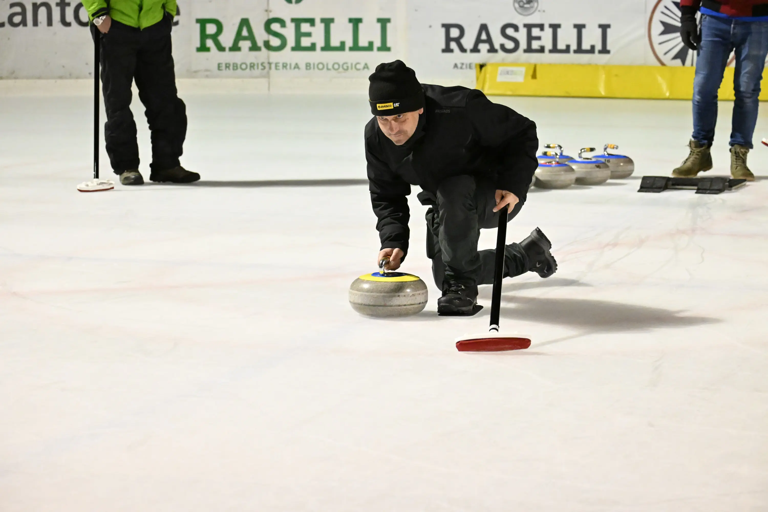 Un giocatore di curling si prepara a lanciare una pietra sul ghiaccio, mentre altri partecipanti osservano. Il giocatore indossa un cappello nero e un giubbotto