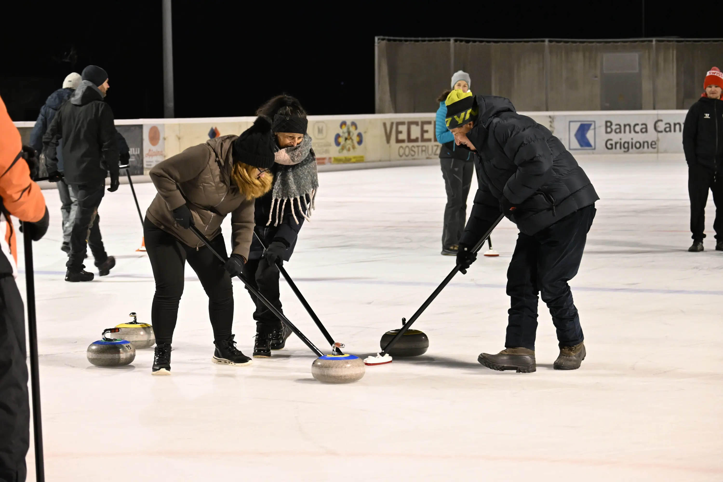 Gruppo di persone che praticano curling su una pista di ghiaccio. Due giocatori si chinano per lanciare delle pietre mentre un terzo osserva. Gli indumenti invern