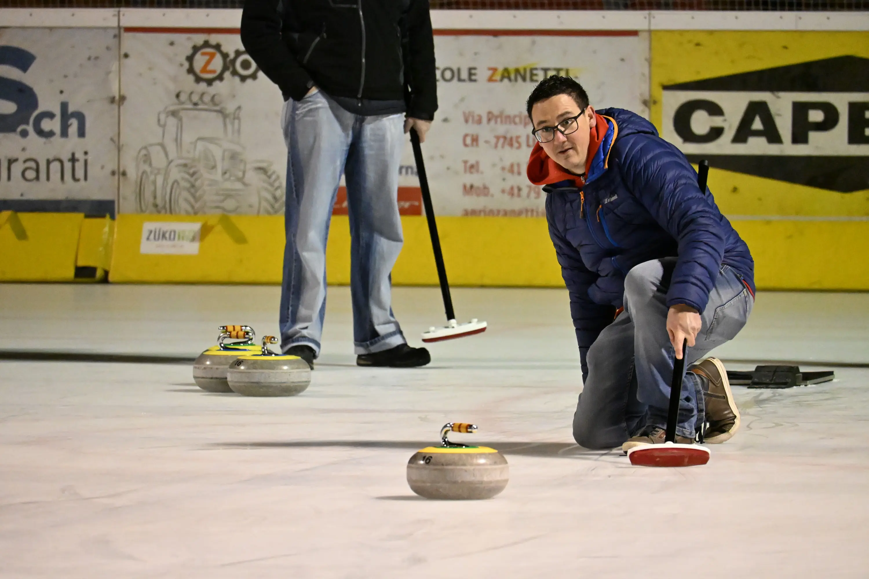 Un giocatore di curling si inginocchia per lanciare una pietra sulla pista, mentre un altro giocatore si trova in piedi sullo sfondo. La superficie di gioco è ghiacci