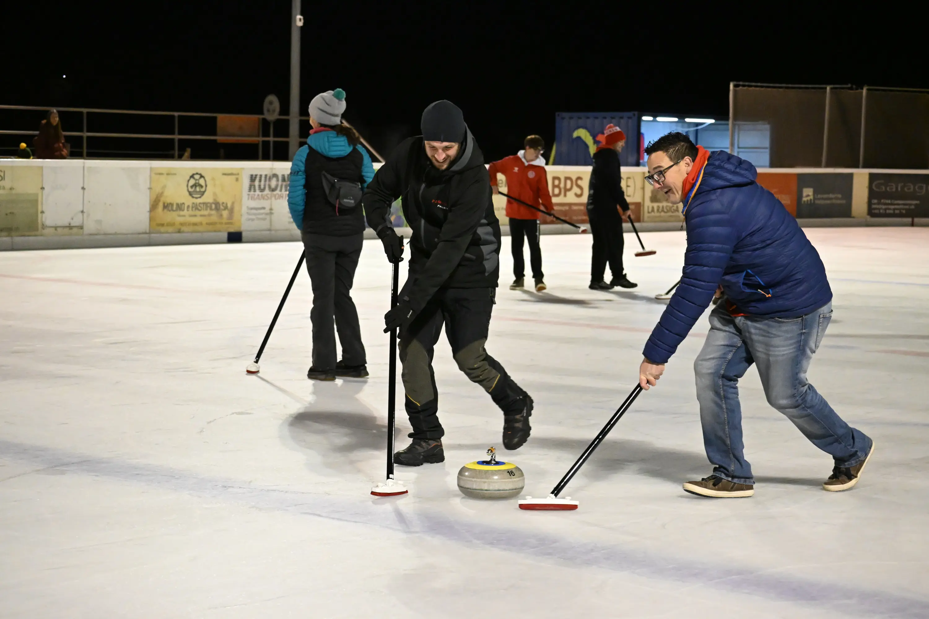 Un gruppo di persone gioca a curling su una pista di ghiaccio all'aperto, con alcuni partecipanti che si preparano a lanciare le pietre e altri che seguono attentamente