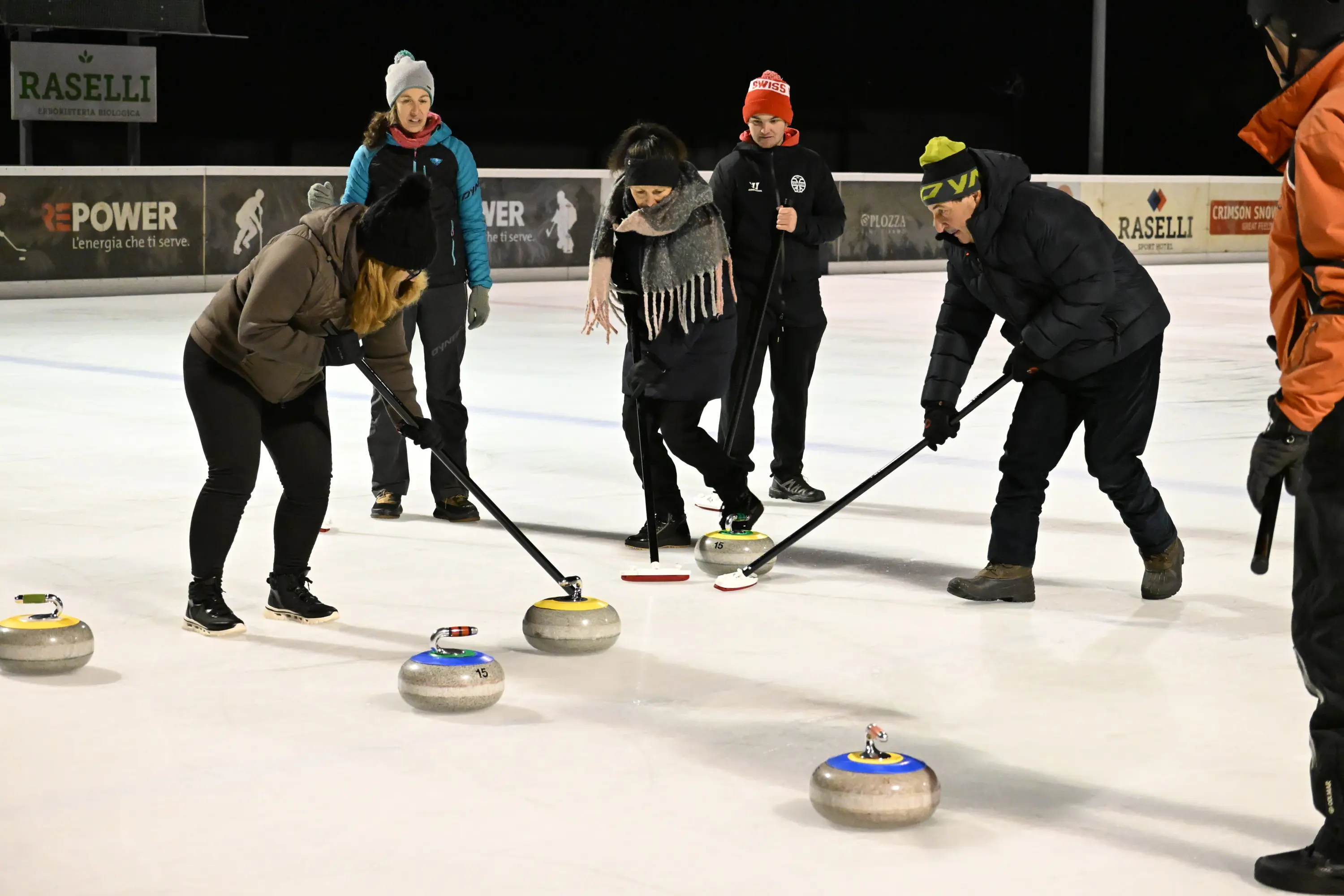 Persone che giocano a curling su un campo di ghiaccio, mentre si piegano per colpire le pietre con le scope. Alcuni indossano giacche e berre