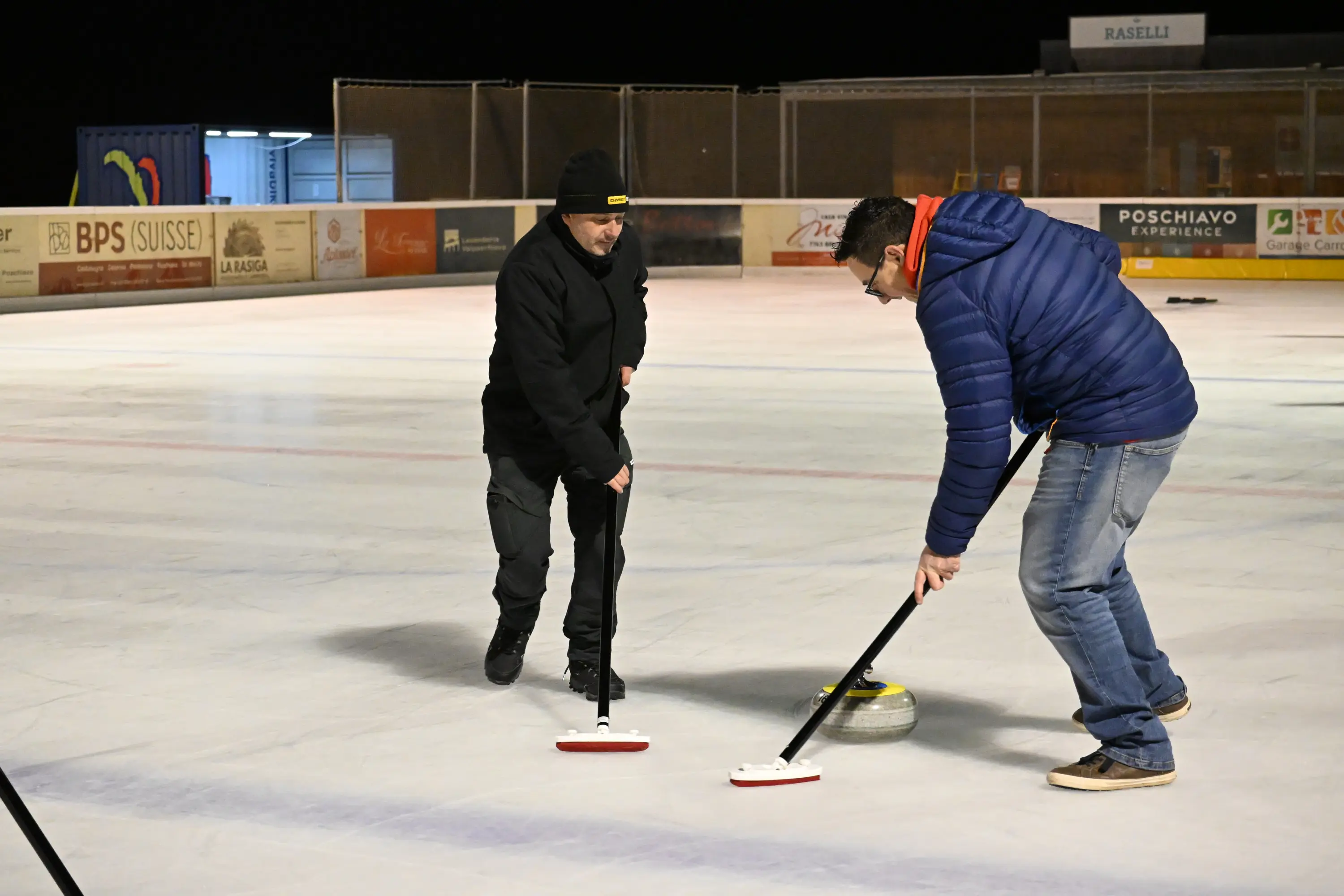 Due uomini su una pista di curling, uno sta lanciando un sasso mentre l'altro lo guida con una scopa. La scena è illuminata, con il ghiaccio visibile e