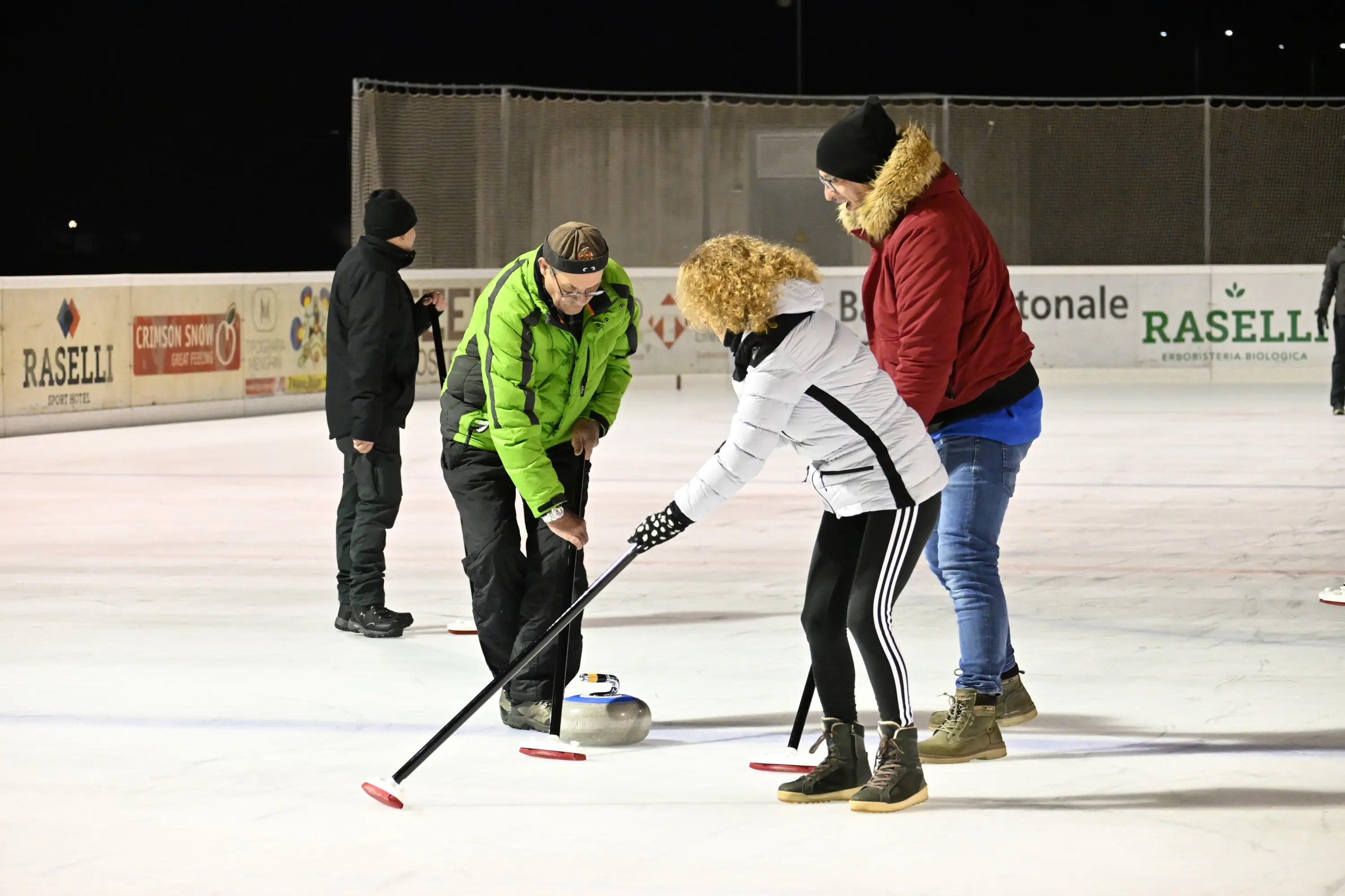 Un gruppo di persone si cimenta nel curling su una pista ghiacciata. Un uomo in giacca verde spiega la tecnica a una donna con giacca bianca e pantaloni a strisce