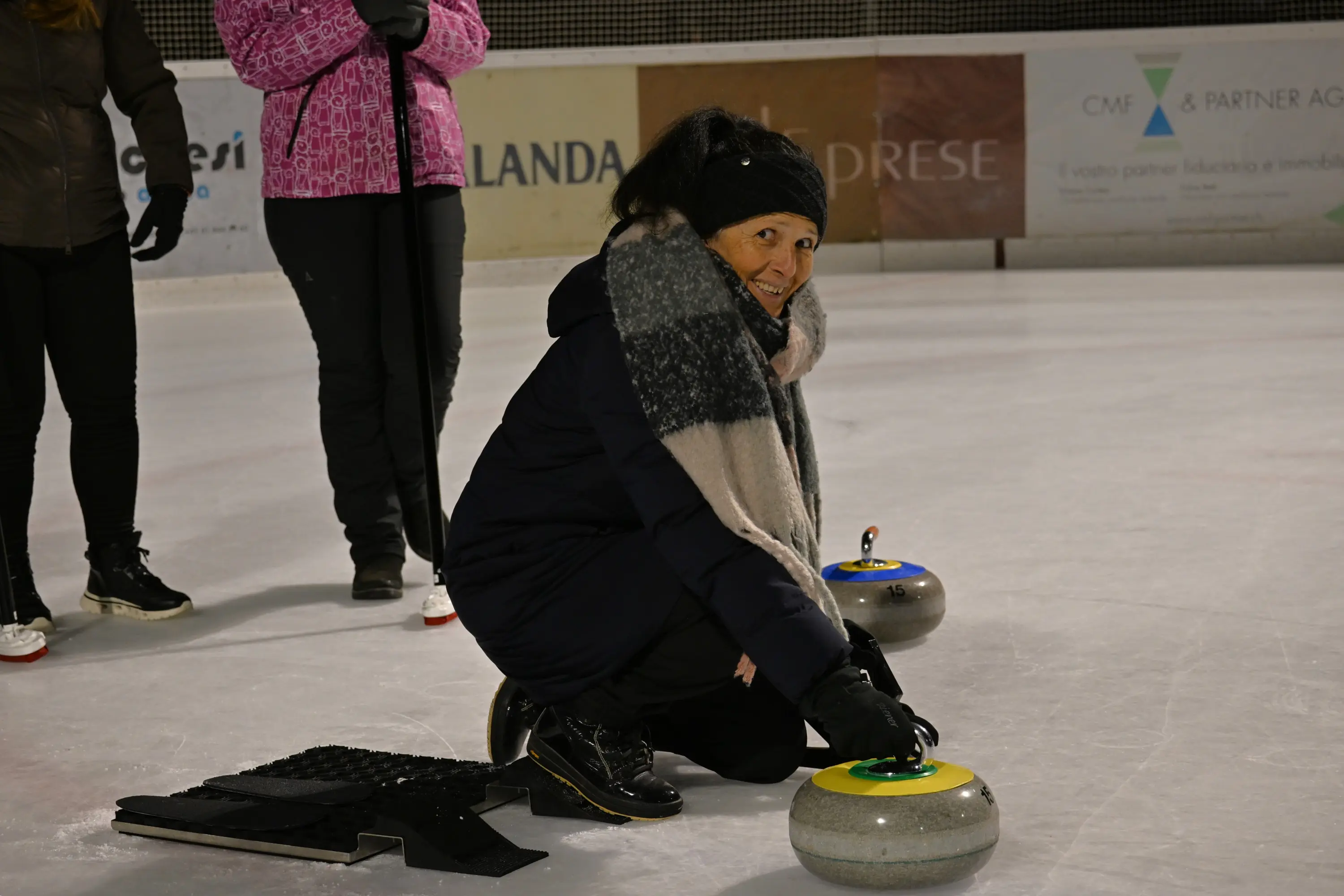 Una donna sorridente si prepara a lanciare una pietra nel curling, inginocchiata sul ghiaccio con una sciarpa e un cappello. Sullo sfondo, altre persone