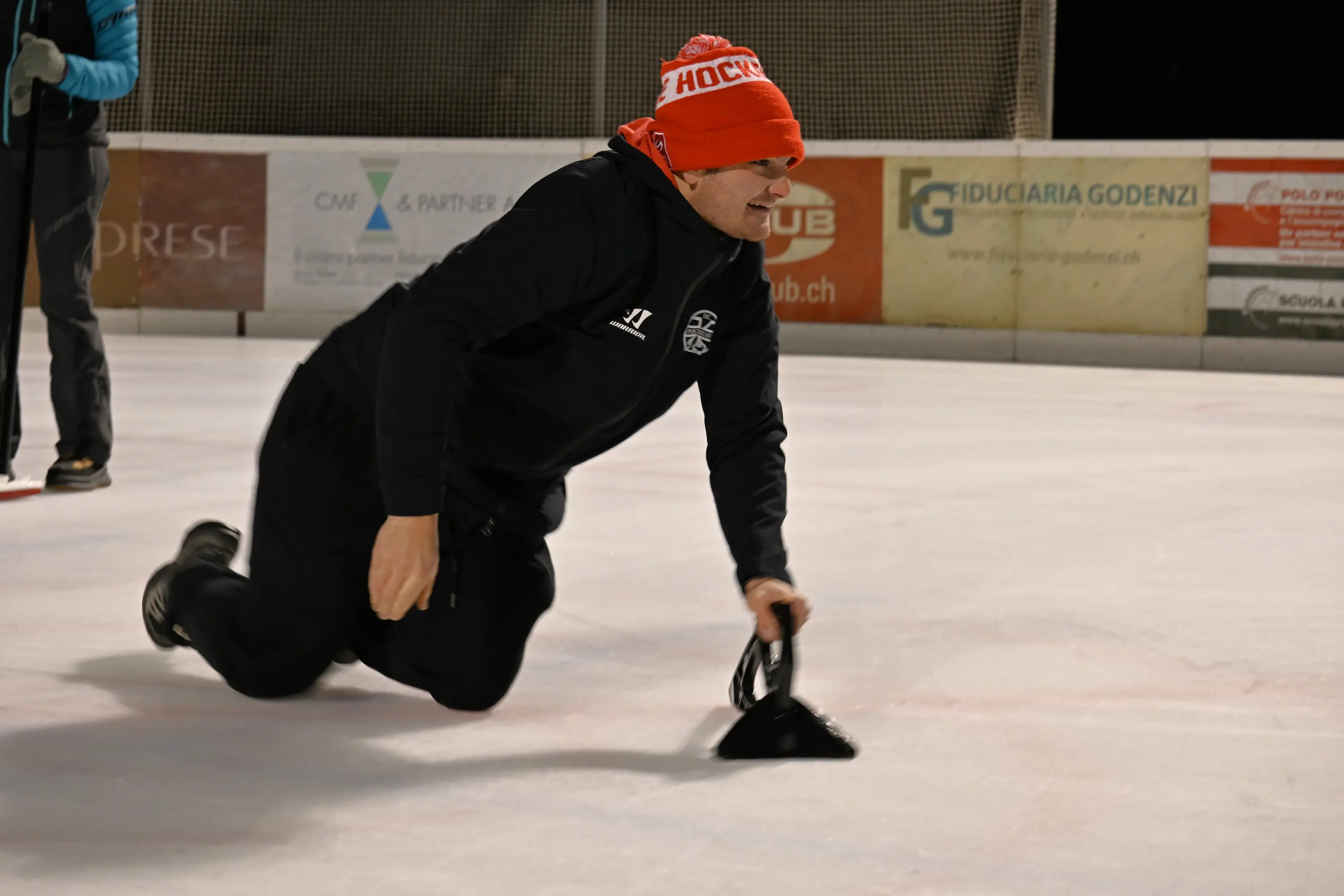 Un uomo in abbigliamento sportivo scivola sulla pista di ghiaccio mentre utilizza uno strumento per pulire la superficie. Indossa un berretto rosso con la scritta