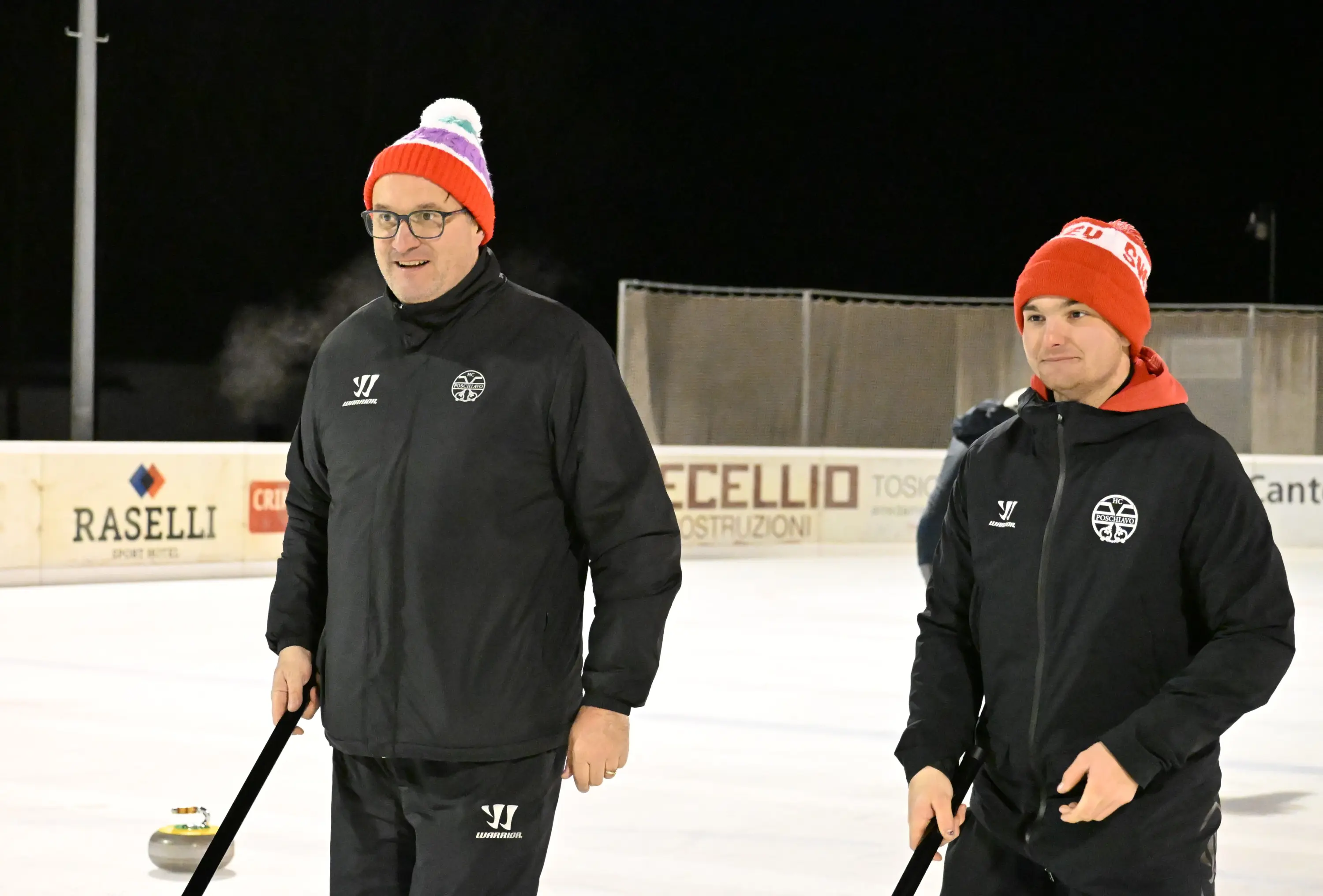 Due uomini sorridenti indossano berretto e abbigliamento sportivo, mentre si preparano a giocare a curling su una pista ghiacciata. Sullo sfondo, si possono