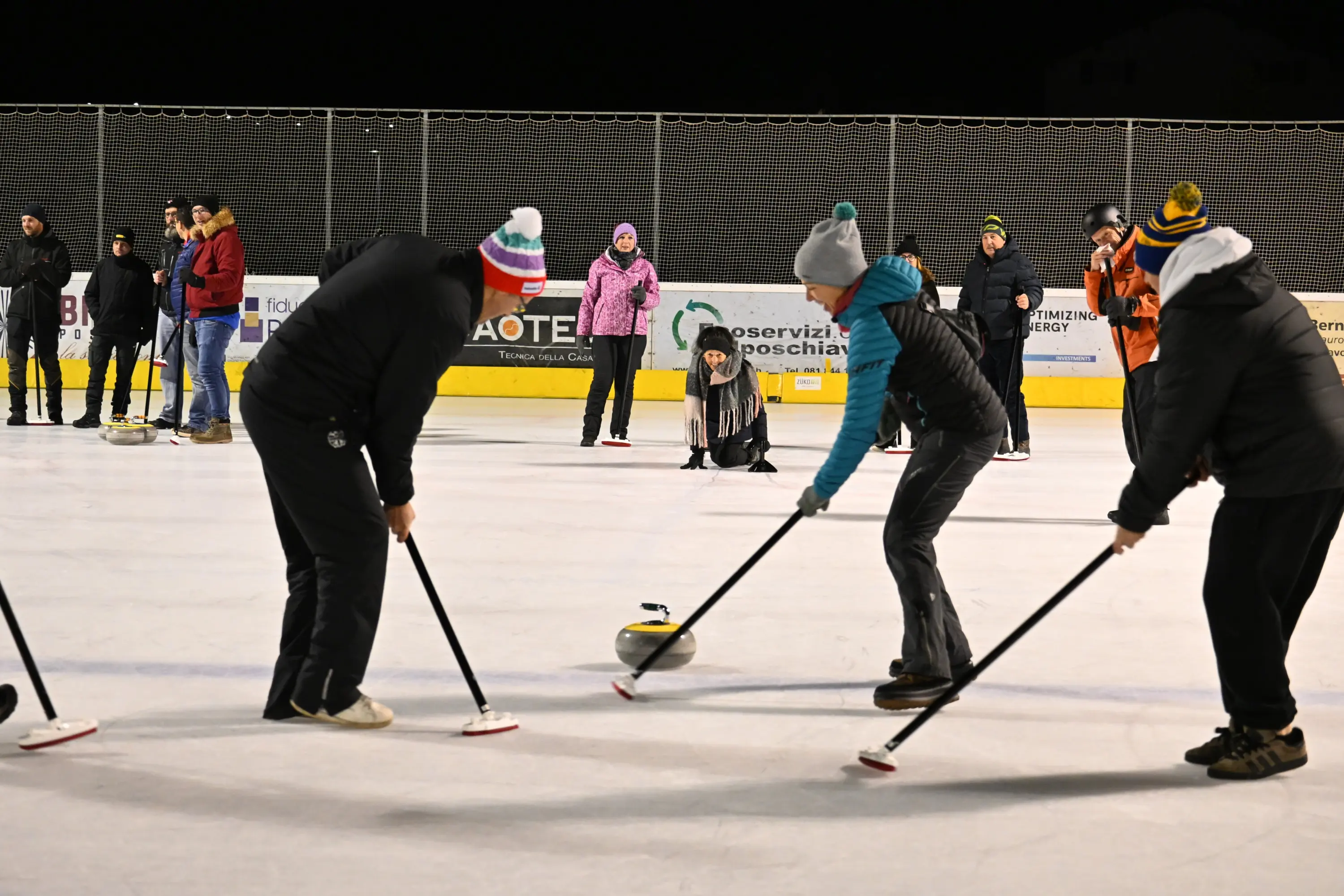 Un gruppo di persone si allena sul ghiaccio, praticando il curling. Alcuni partecipanti si piegano per colpire il stone, mentre altri osservano. È una serata