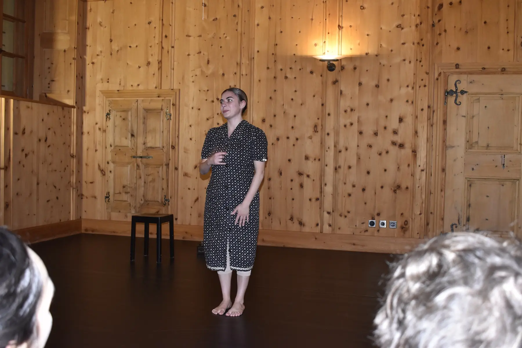 Una persona in abbigliamento scuro a pois esegue una performance in una stanza con pareti di legno. Si trova su un pavimento scuro, di fronte a un pubblico parzialmente visibile.