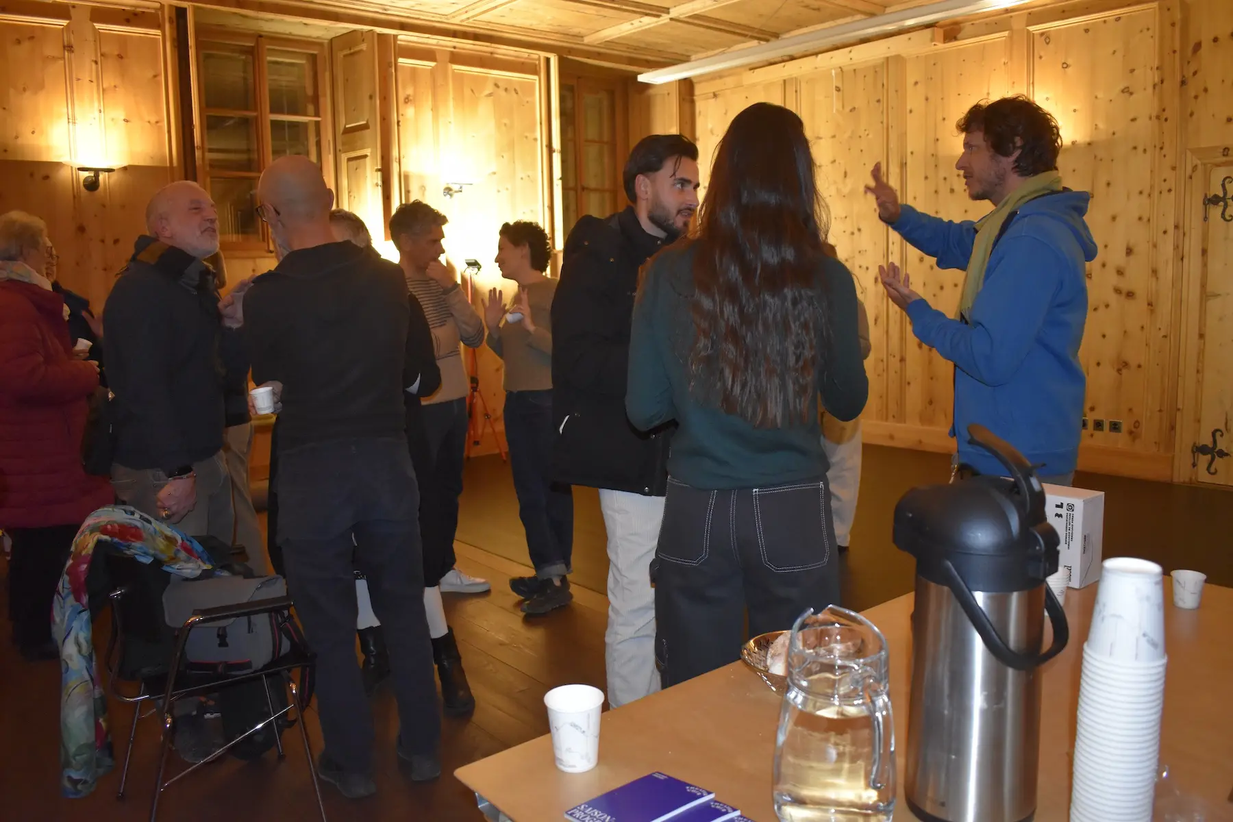 Un gruppo di persone si trova in una stanza in legno, conversando e socializzando. Alcuni partecipanti stanno bevendo da bicchieri di carta. In primo piano, una persona in blu sta parlando animatamente con un altro, mentre altre listen attentamente. Su un tav