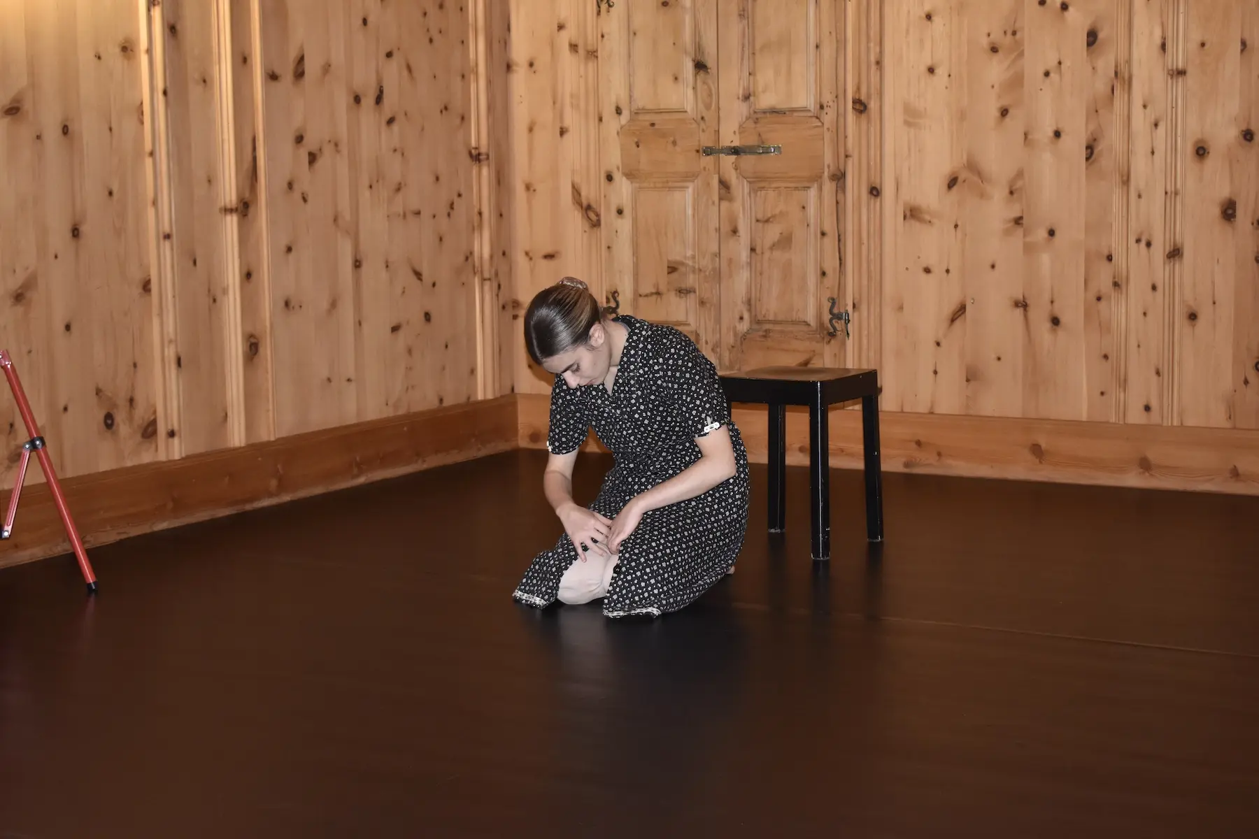 Una donna in abito a pois, in ginocchio su un pavimento nero, sembra riflettere o danzare in una stanza con pareti di legno. Dietro di lei c'è una sedia nera.