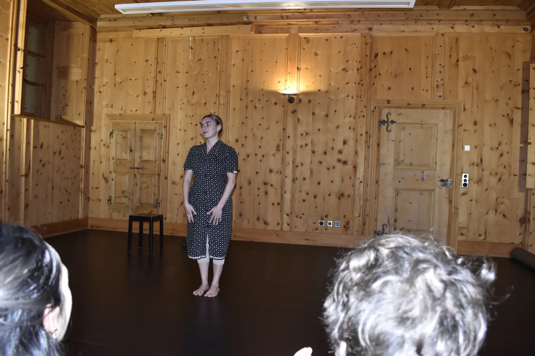 Una donna in abiti scuri si trova al centro di una stanza con pareti di legno chiaro. Sta eseguendo un'esibizione, mentre il pubblico, visibile in primo piano, la osserva attentamente. Un piccolo sgabello nero è posizionato accanto