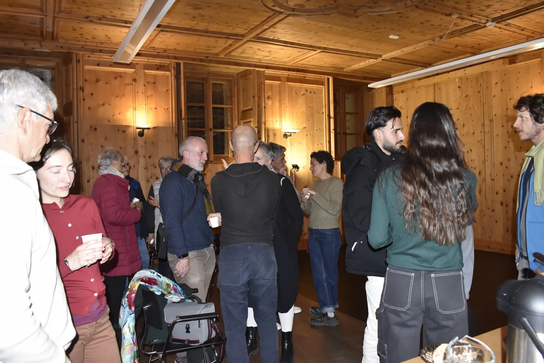 Un gruppo di persone si riunisce all'interno di una sala con pareti di legno. Alcuni conversano mentre altri sorseggiano da bicchieri. L'atmosfera è accogliente e informale.