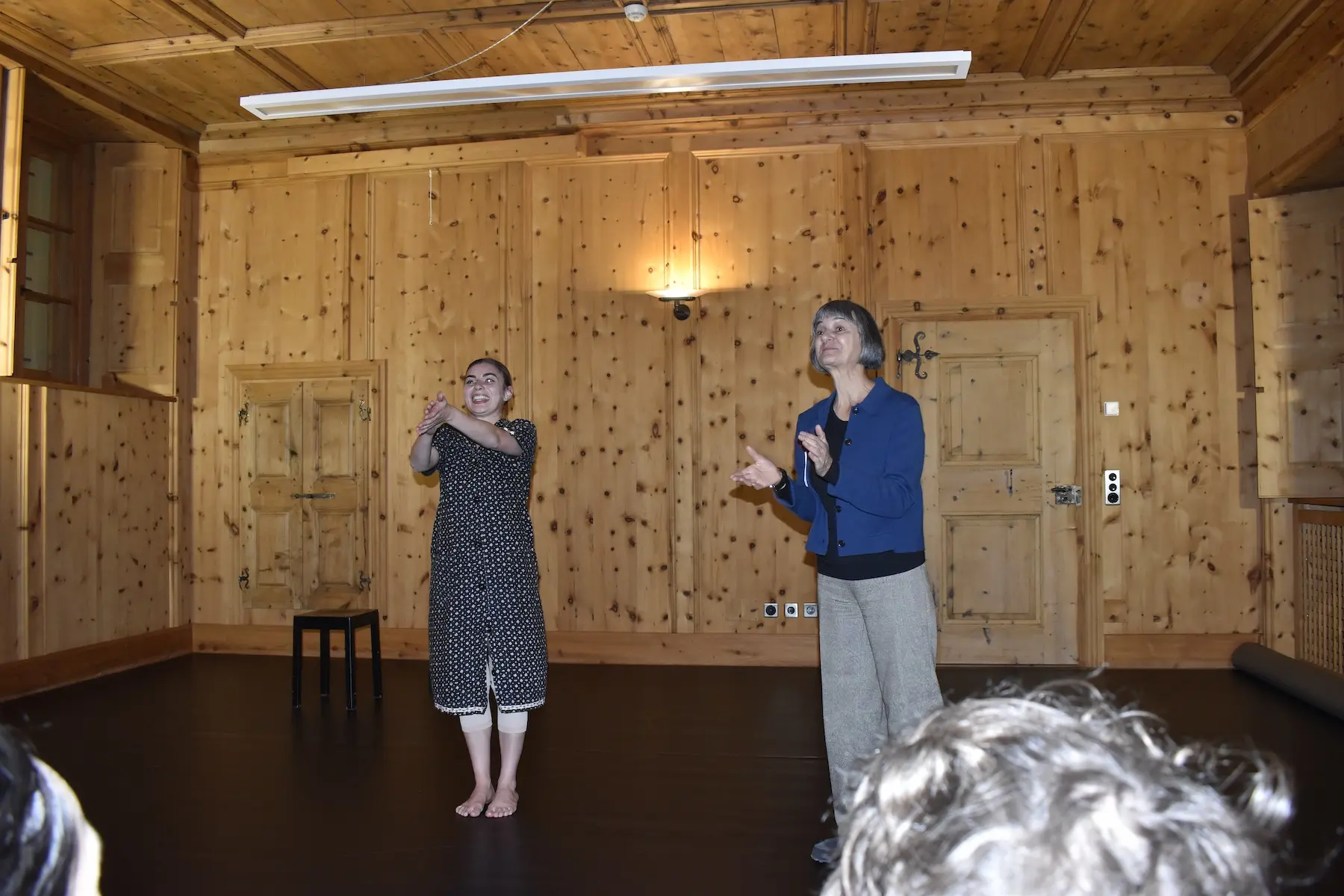 Due donne si esibiscono in una stanza con pareti di legno. Una indossa un vestito a pois e sta gesticolando, mentre l'altra, in abbigliamento blu, applaude. Un semplice sgabello nero si trova accanto a loro.