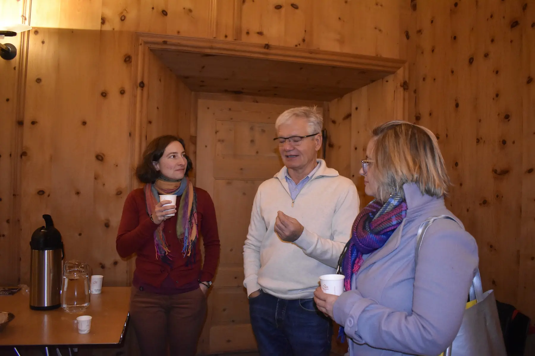 Due persone con tazze in mano ascoltano un uomo che parla, all'interno di una stanza con pareti di legno. Tutti sorridono e indossano sciarpe colorate.