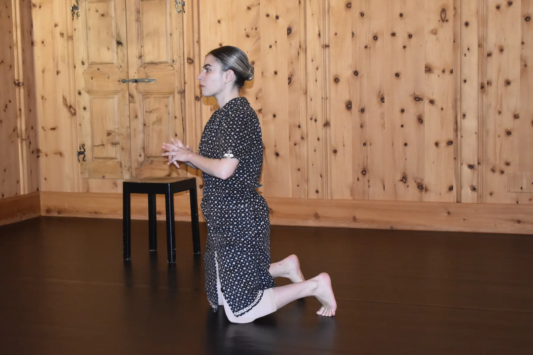 Una giovane donna è inginocchiata sul pavimento di legno scuro, con le mani unite in preghiera. Indossa un abito a pois e si trova in una stanza con pareti di legno chiaro. Un piccolo sgabello nero è visibile sullo sf