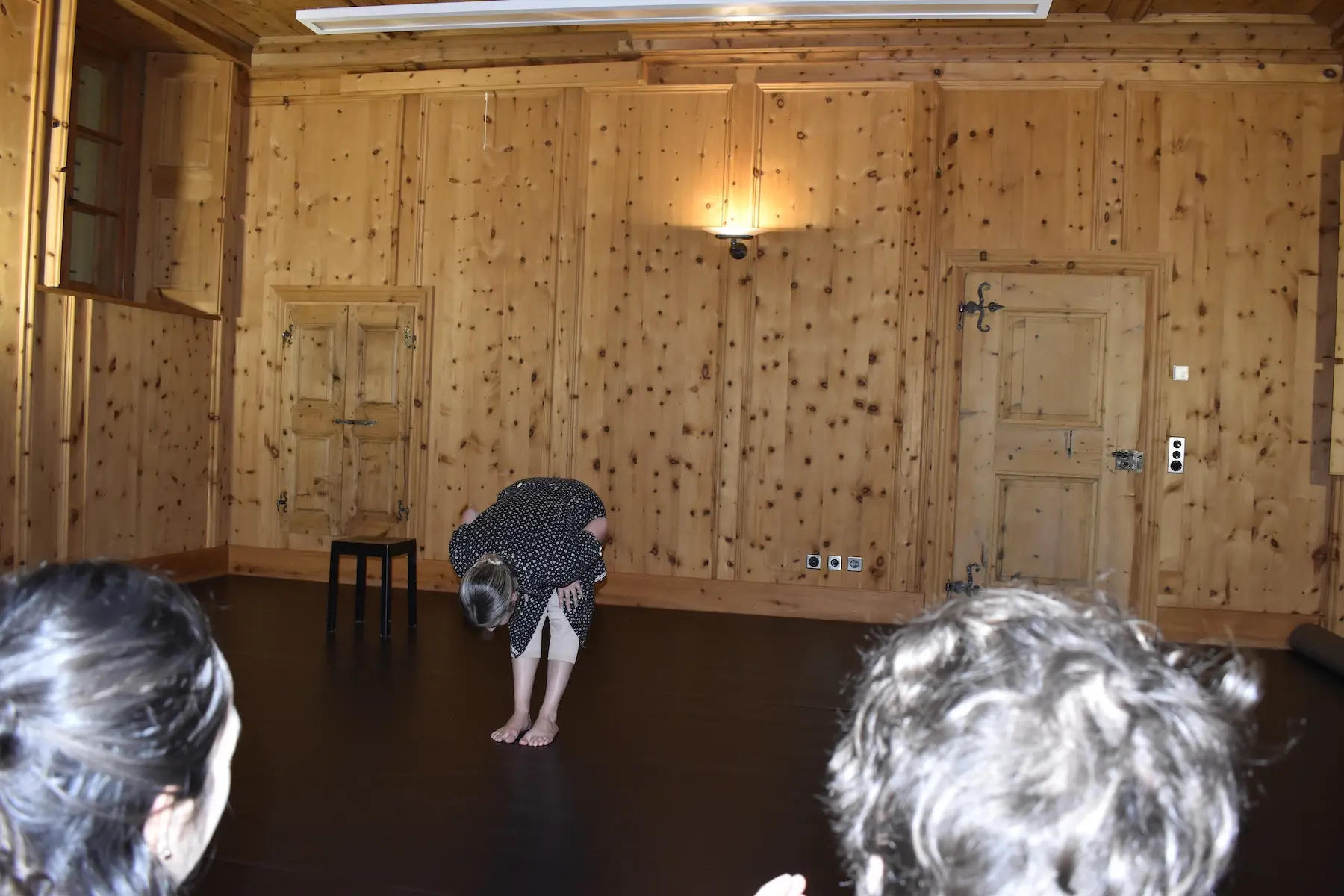 Una donna si inclina in avanti su un palco vuoto, circondata da pareti di legno. In primo piano, si vedono le spalle di due persone che osservano.