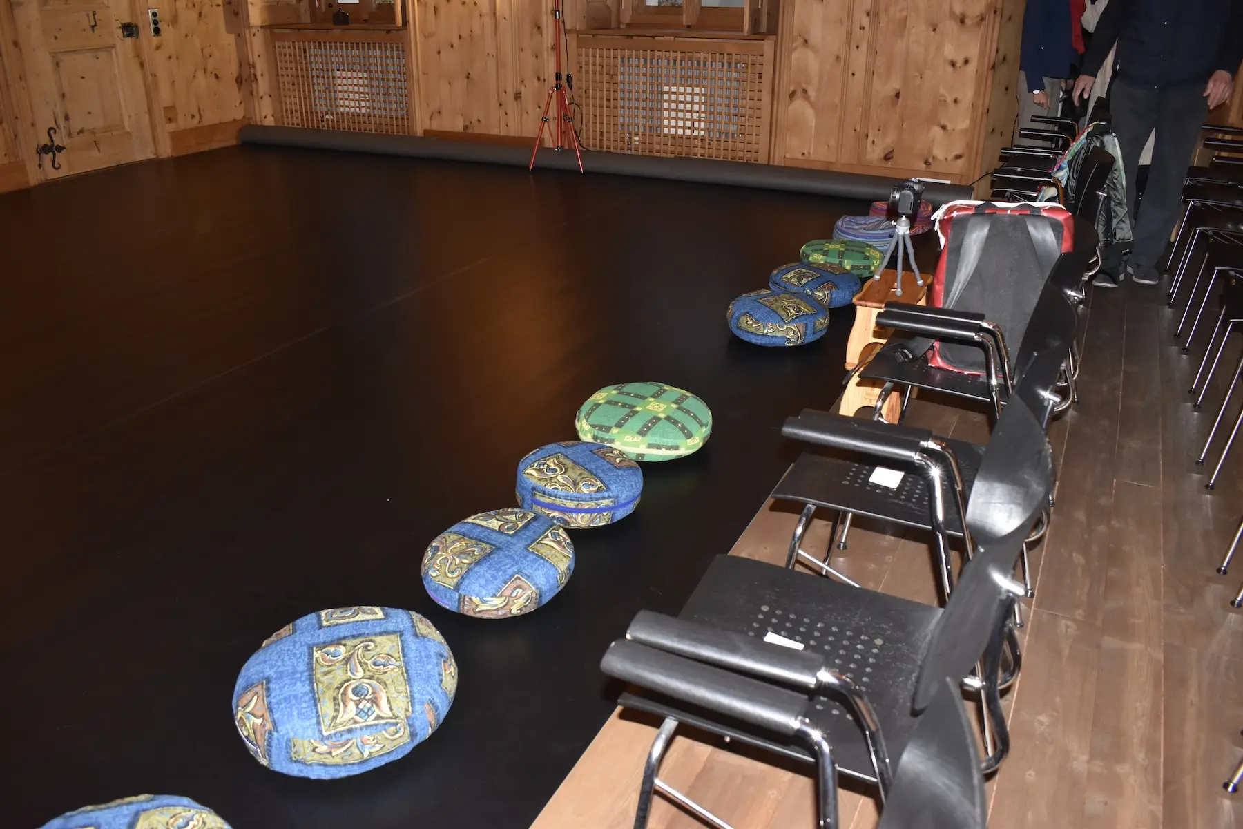 Un pavimento di legno scuro con cuscini colorati disposti in fila, accanto a delle sedie nere. L'ambiente ha pareti in legno e finestre in alto.