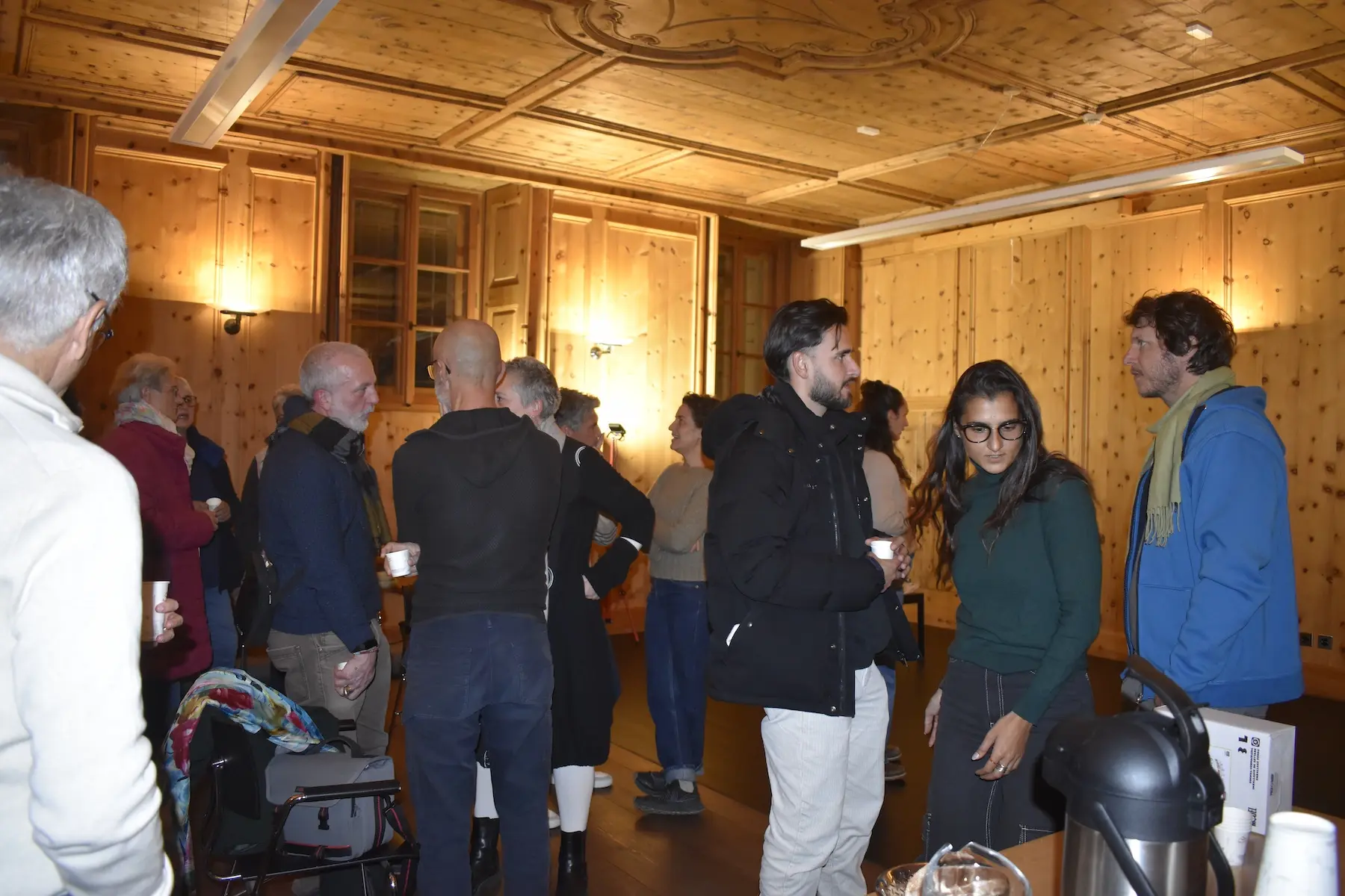 Un gruppo di persone conversano all'interno di una sala in legno, con luci soffuse e un'atmosfera accogliente. Alcuni partecipanti tengono in mano delle tazze.