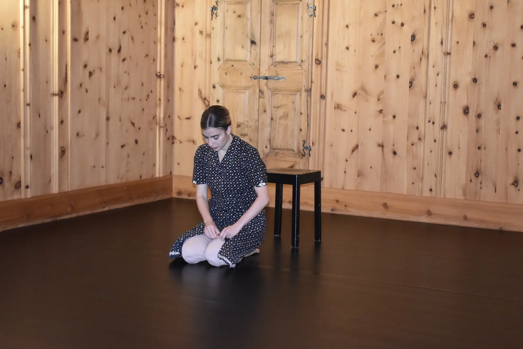 Una giovane donna in abito a pois si trova inginocchiata su un pavimento scuro, mentre si aggiusta le calze. È in una stanza con pareti di legno chiaro e una porta chiusa sullo sfondo. C'è una sedia nera accanto