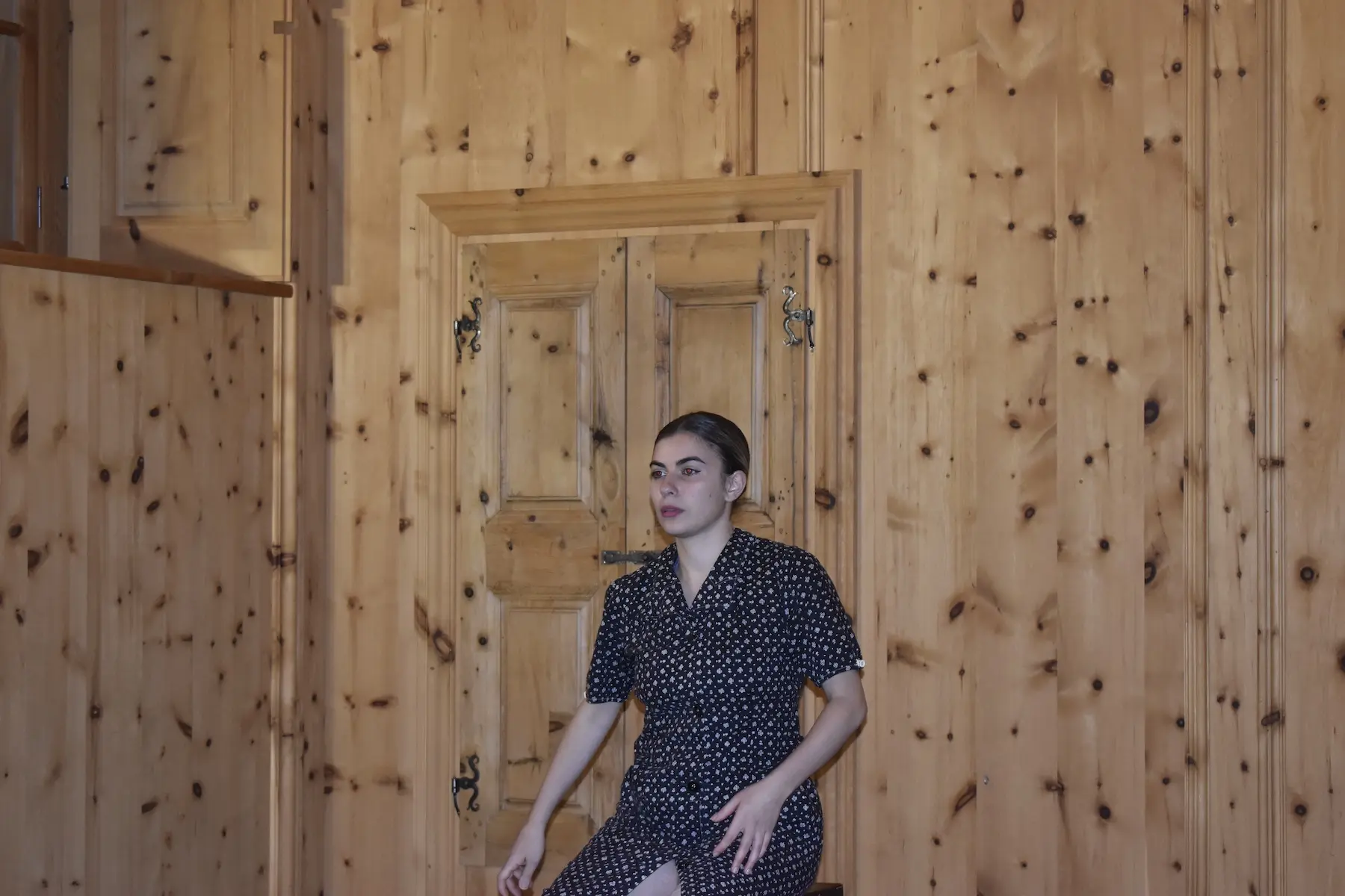 Una giovane donna seduta su una sedia in una stanza con pareti di legno. Indossa un vestito nero a pois bianchi e ha i capelli raccolti. Dietro di lei c'è una porta in legno con maniglie decorative.