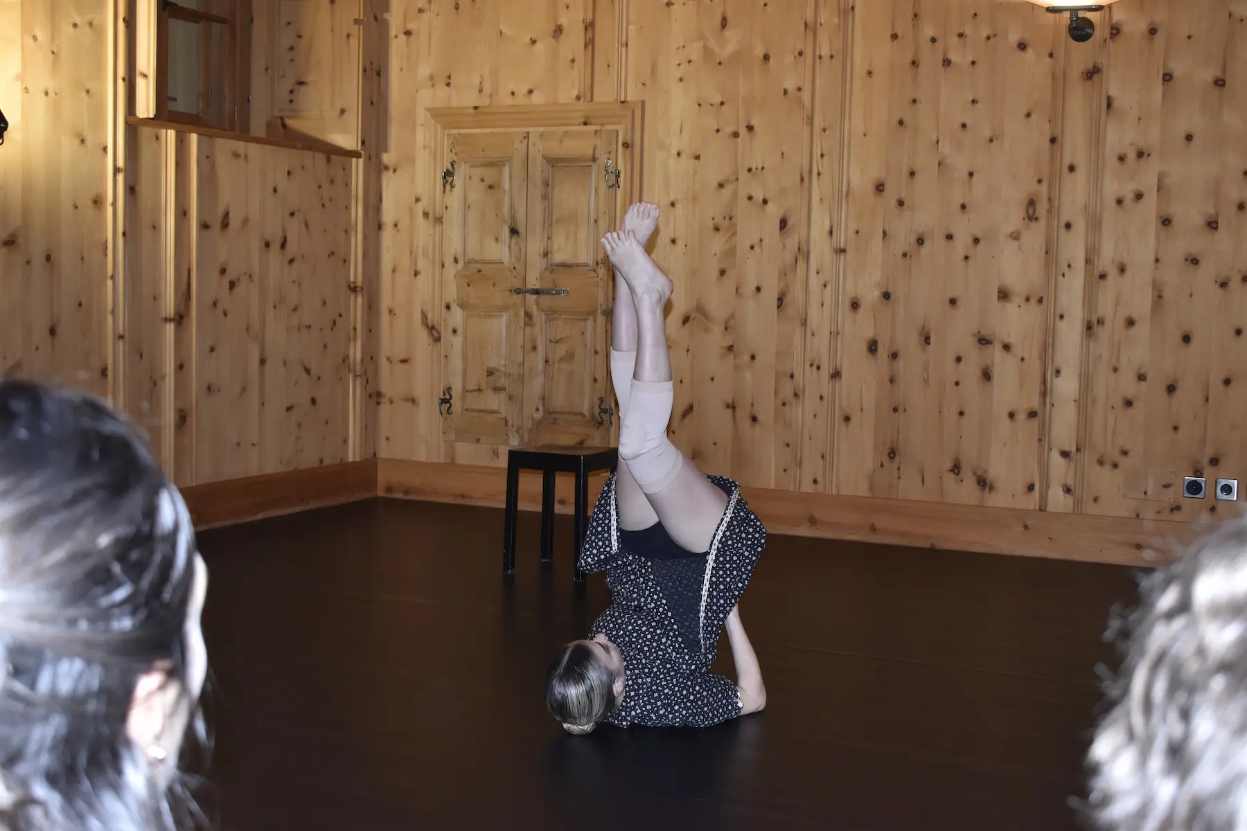 Una ballerina esegue un movimento sul pavimento, con le gambe sollevate verso l'alto. La stanza ha pareti di legno e un'atmosfera accogliente. Un paio di persone sono visibili in primo piano, rivolte verso la ballerina.