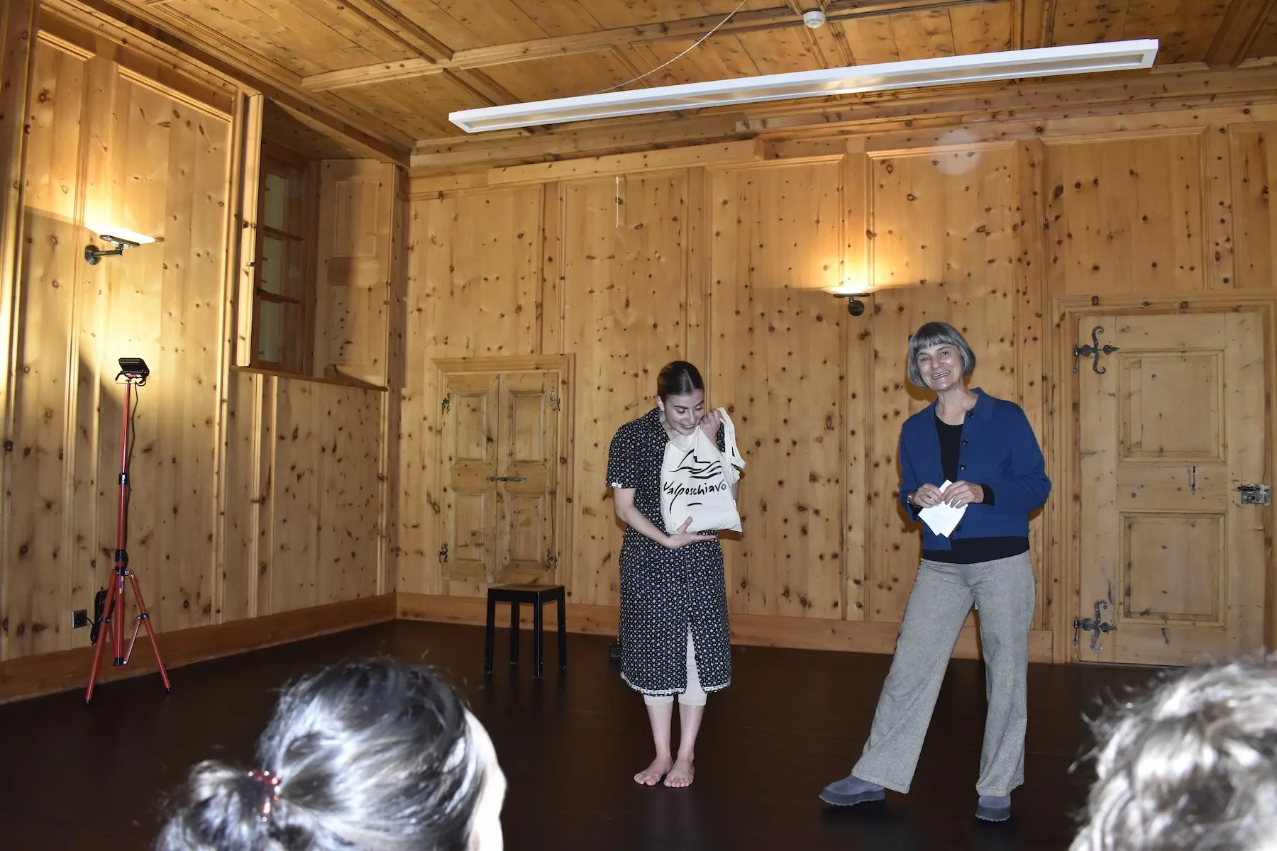 Due donne si trovano in una stanza con pareti in legno. Una tiene in mano un oggetto, mentre l'altra sorride. Una sedia nera è visibile sullo sfondo. Illuminazione calda e accogliente.