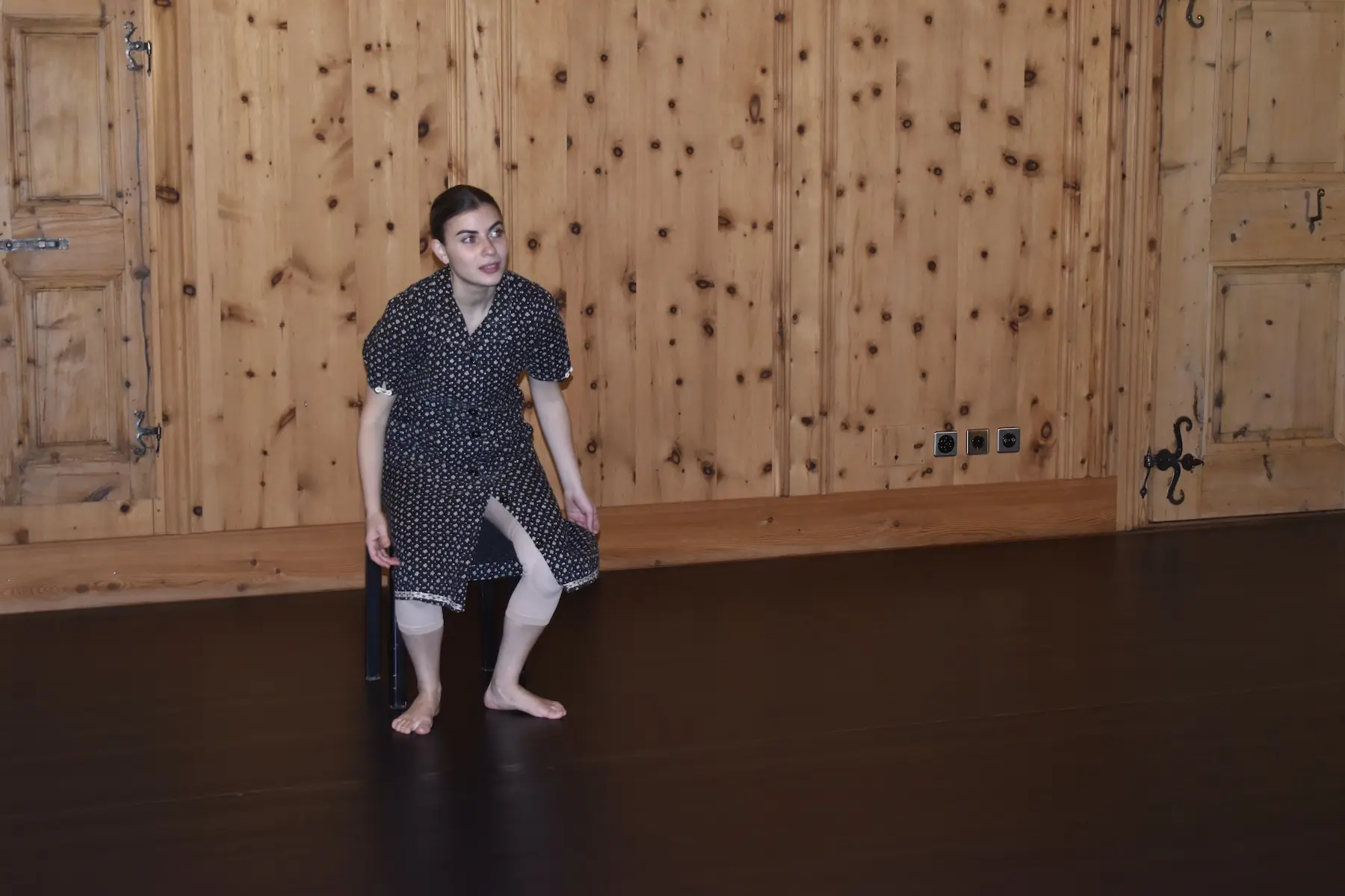 Una ballerina in abito a pois esegue una danza su un pavimento scuro, sullo sfondo di una parete di legno.