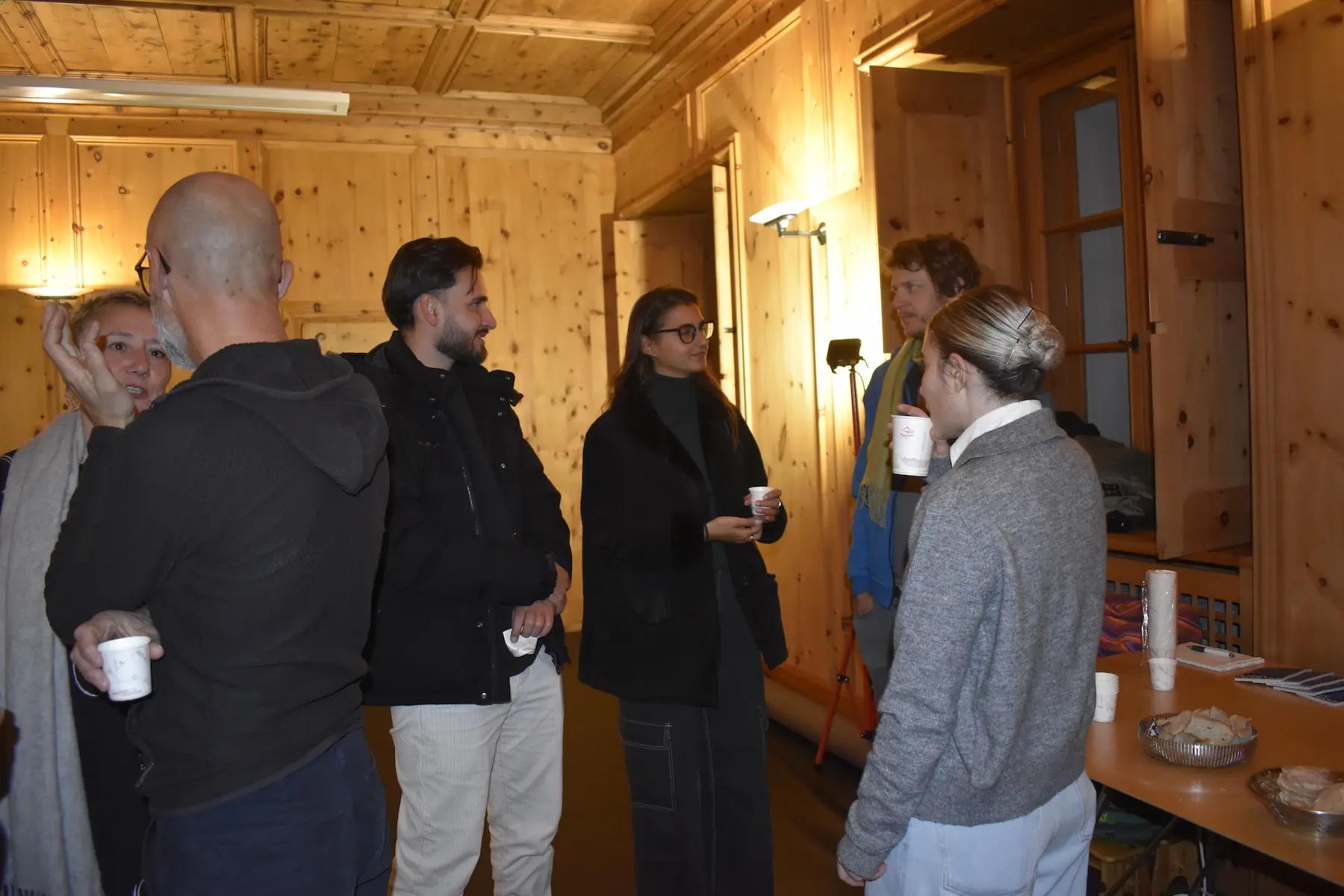 Un gruppo di persone si trova in una stanza con pareti di legno. Alcuni stanno conversando, mentre altri tengono delle tazze in mano. L'atmosfera è informale e conviviale.