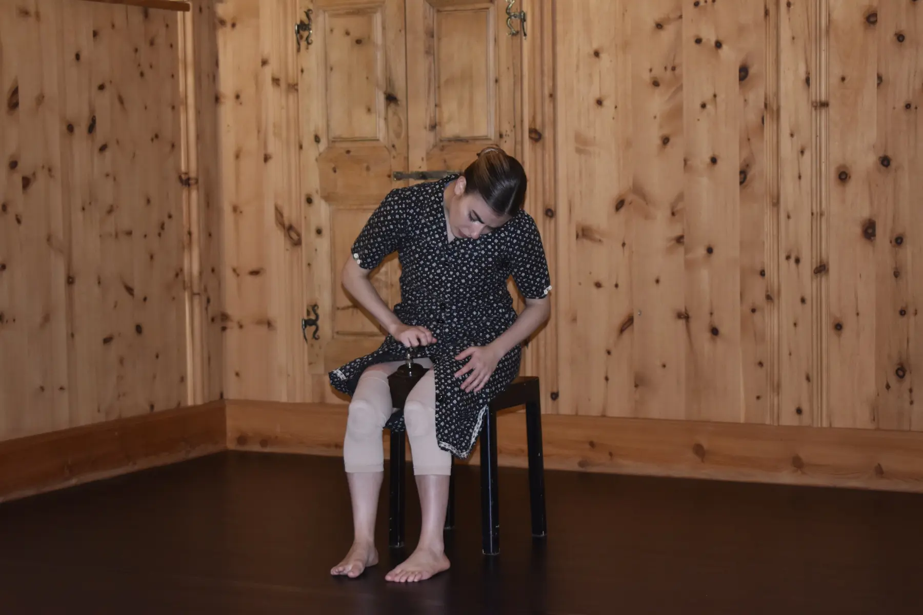 Una donna seduta su una sedia nera in una stanza con pareti di legno. Indossa un abito a pois e sembra concentrata su un oggetto tra le mani. I piedi nudi poggiano sul pavimento scuro.
