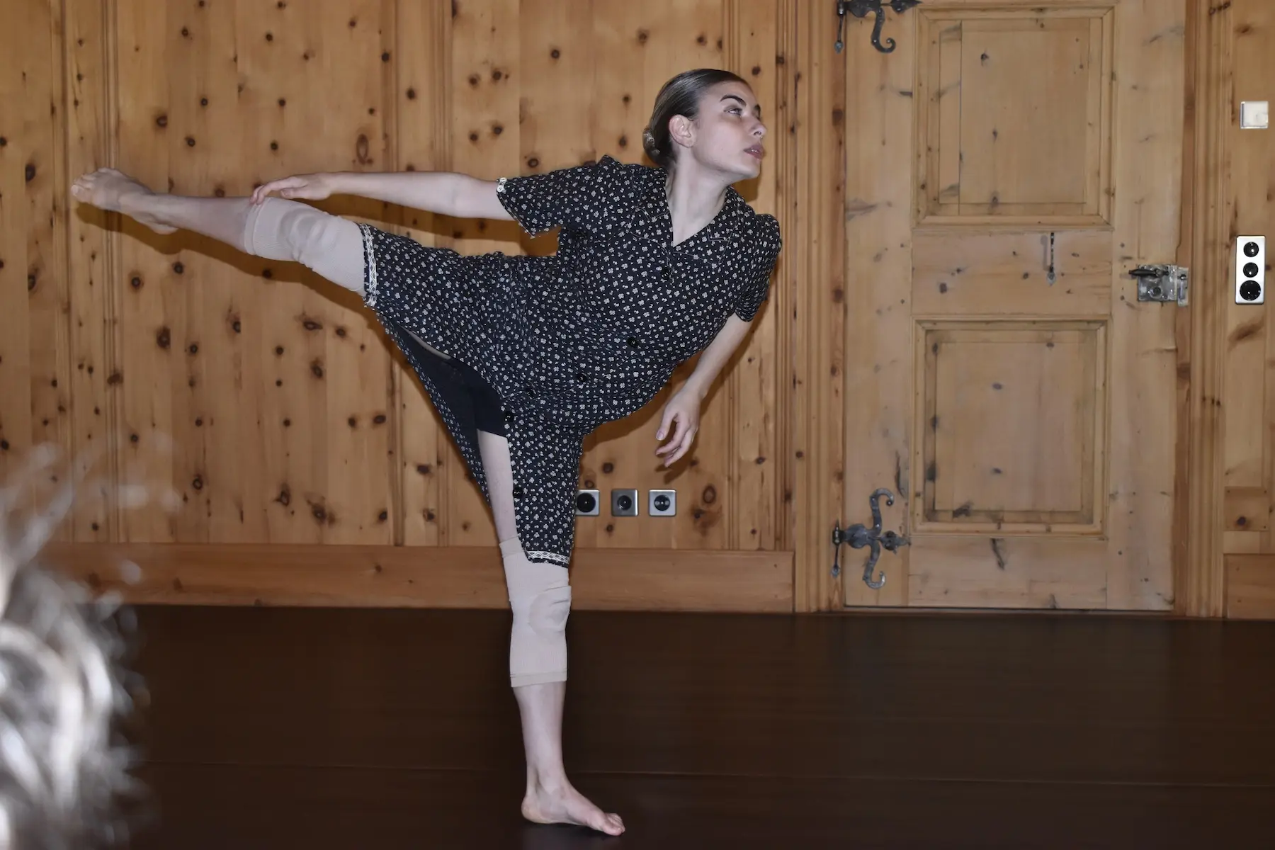 Una ballerina in abito nero con pois esegue una posa elegante, sollevando una gamba mentre si inclina verso il lato. Il fondo è di legno chiaro, creando un ambiente caldo e naturale.