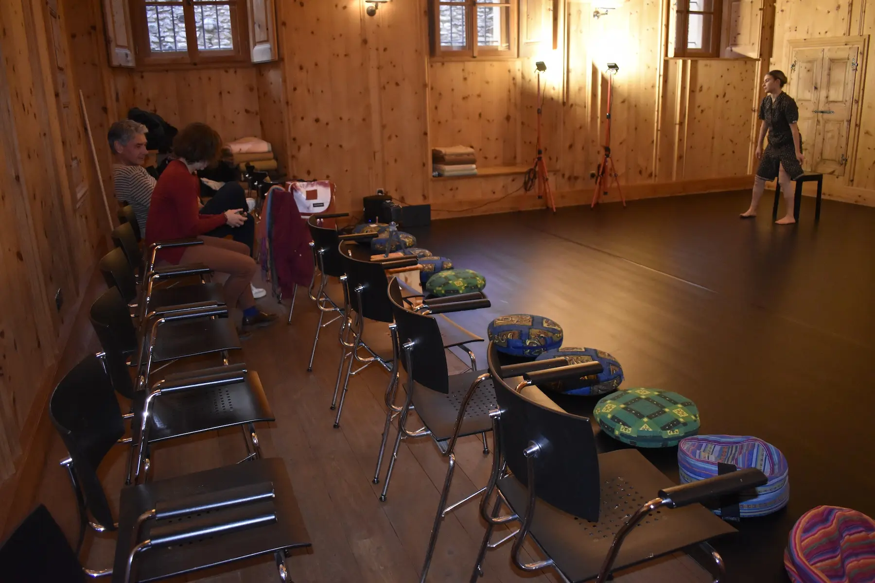Una sala con pareti in legno e un pavimento scuro. Si vedono sedie disposte in fila e cuscini colorati sui sedili. Due persone sono sedute a sinistra, mentre una donna si sta esibendo in piedi a destra. Illuminazione cal