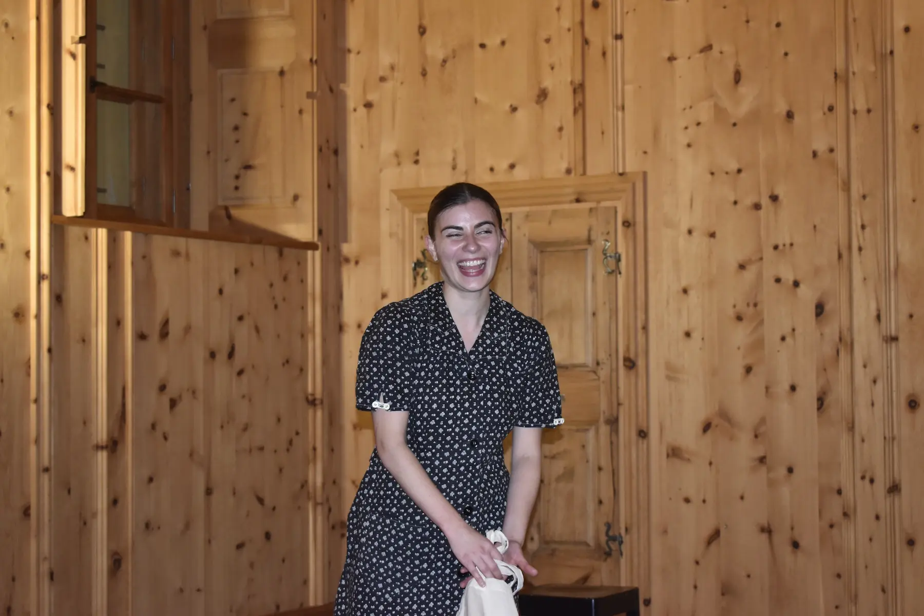 Una giovane donna sorride mentre si trova in una stanza con pareti di legno. Indossa un vestito a pois e tiene in mano un oggetto bianco.