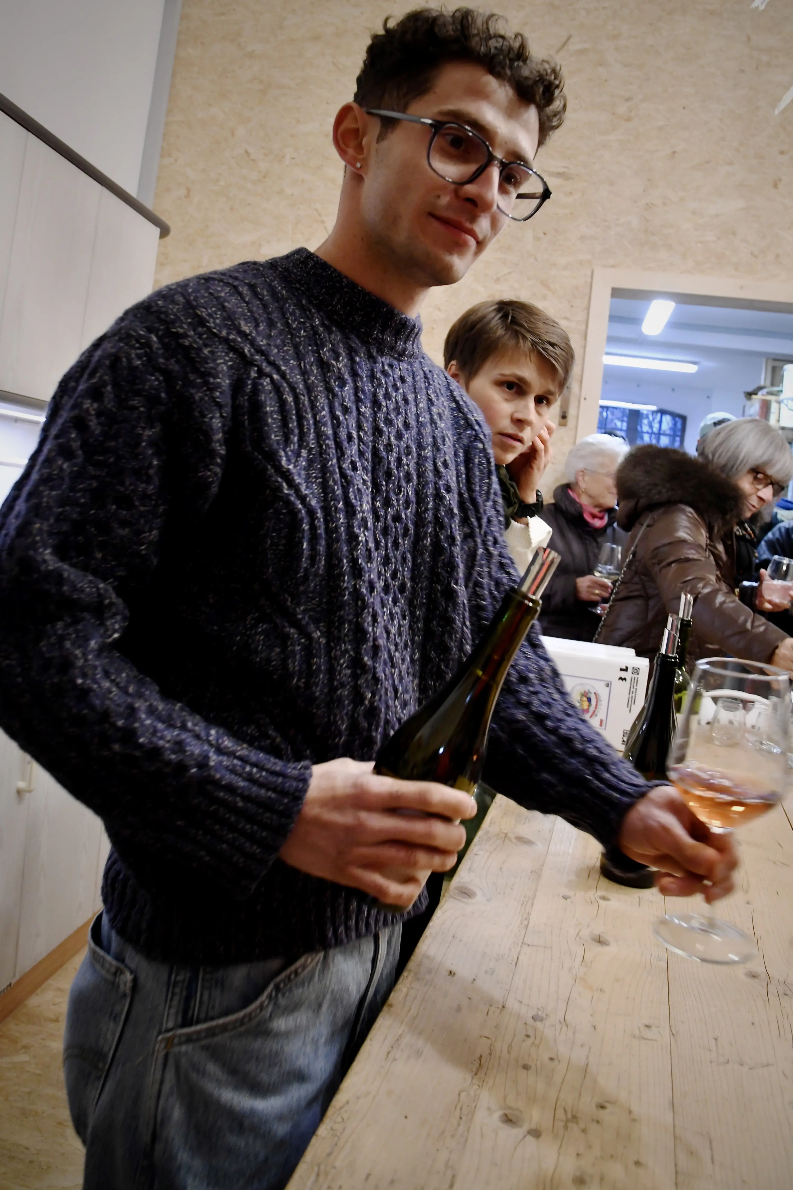 Un giovane con occhiali indossa un maglione blu a coste e tiene in mano una bottiglia di vino e un bicchiere. Sullo sfondo, un'altra persona osserva con interesse.